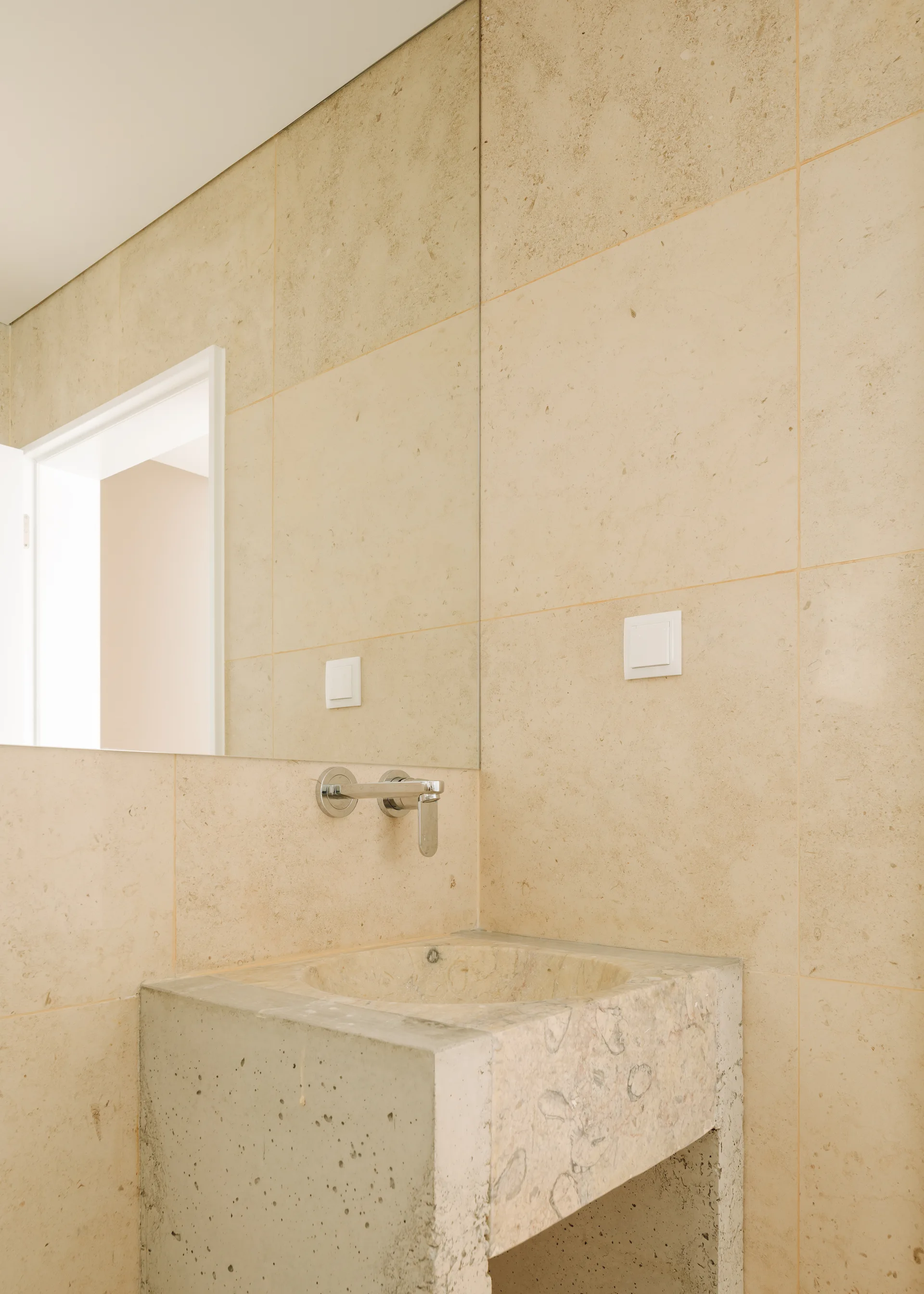 A minimalist bathroom features a textured concrete washbasin under a wall-mounted metal faucet against a wall of large beige stone tiles.