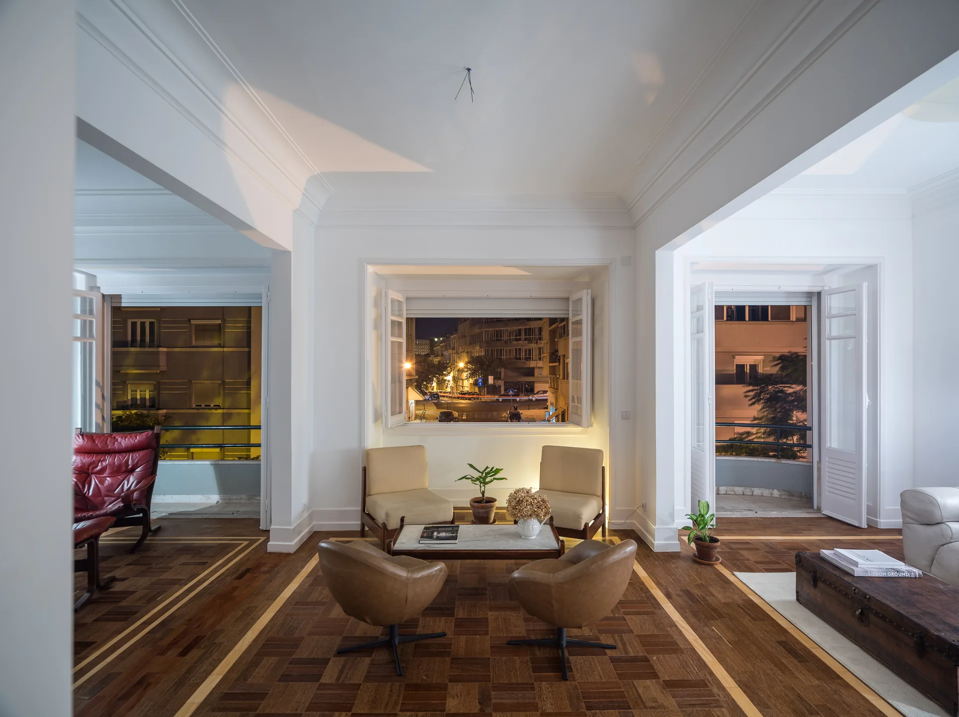 A white apartment living room with patterned parquet floors, tan armchairs, a marble coffee table, and an open window showing a city street at night.