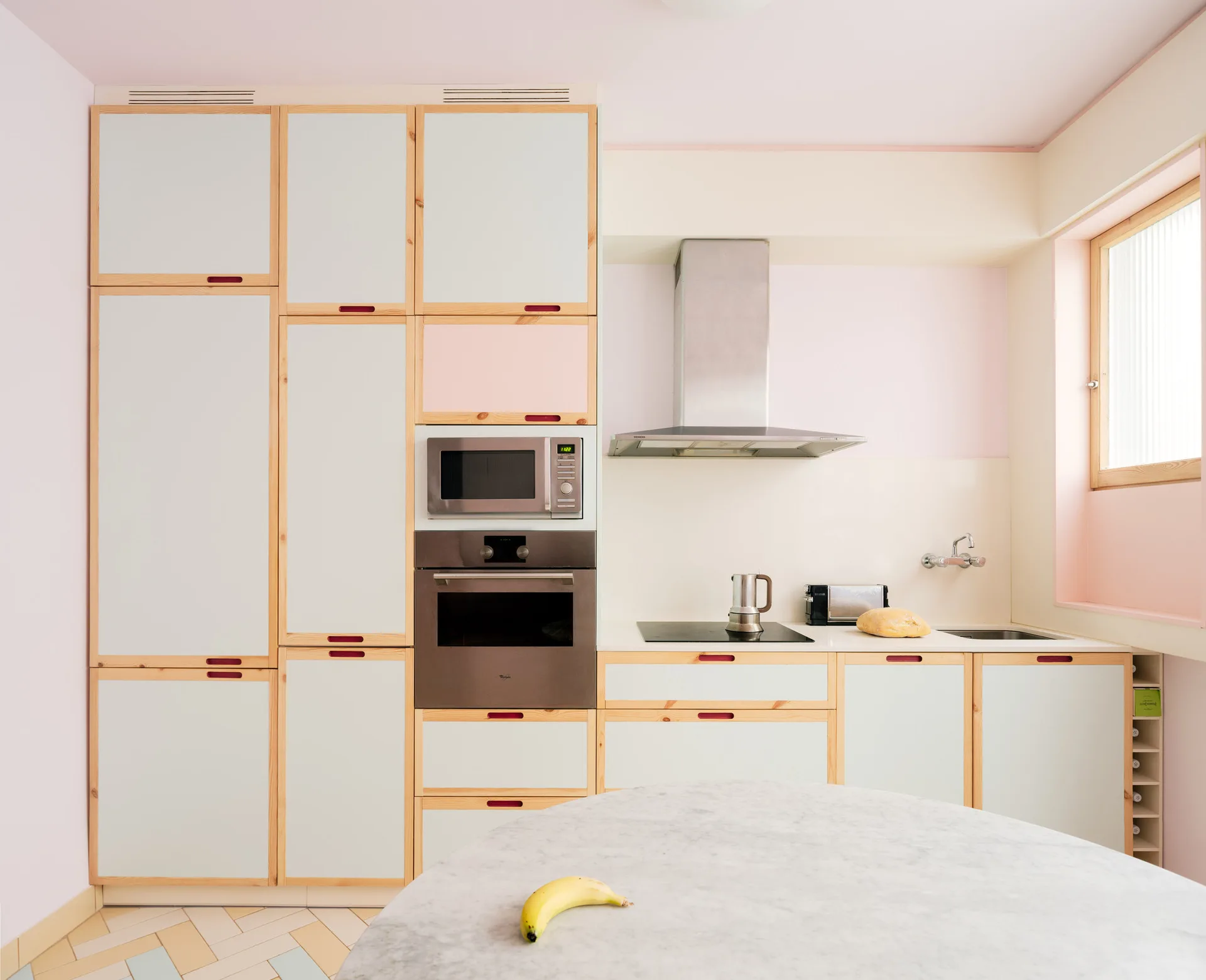 A bright kitchen features light wood-framed cabinetry, a stainless steel oven and range hood, pale pink walls, and a round marble table.