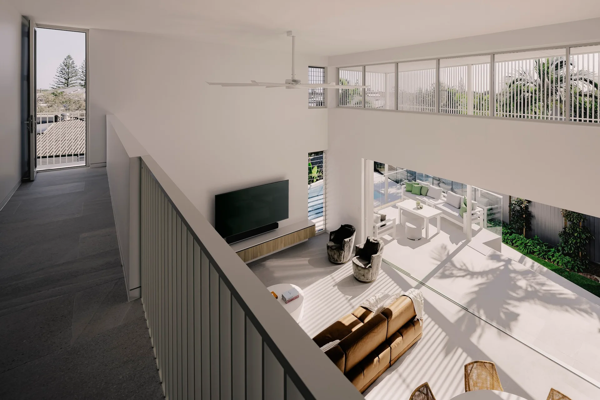 A high-angle view looking down from a mezzanine at a modern white living room with a tan leather sofa and large glass doors leading outside.