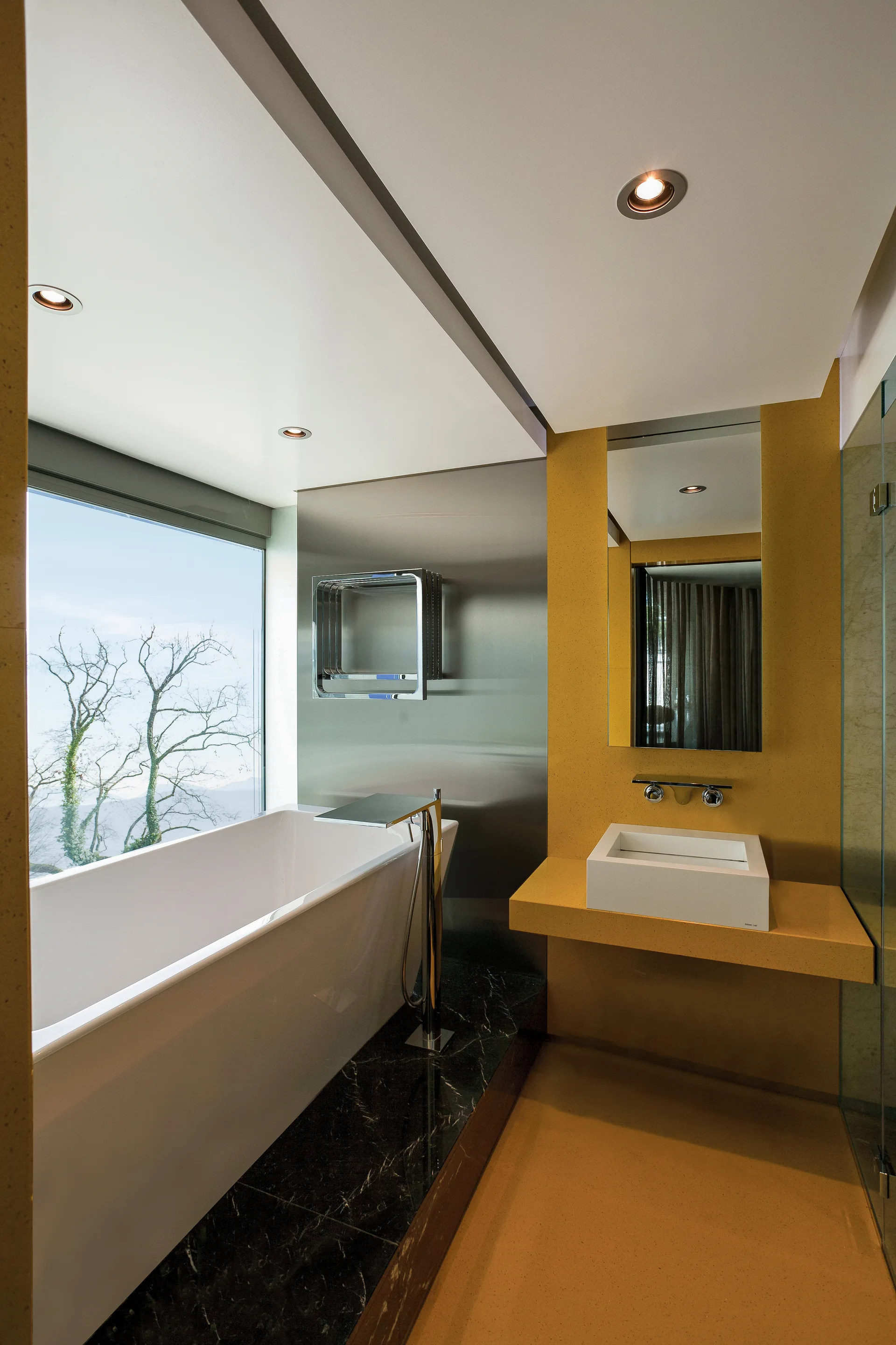 A modern bathroom features a white freestanding bathtub on a dark marble floor next to a mustard yellow wall with a floating vanity sink.