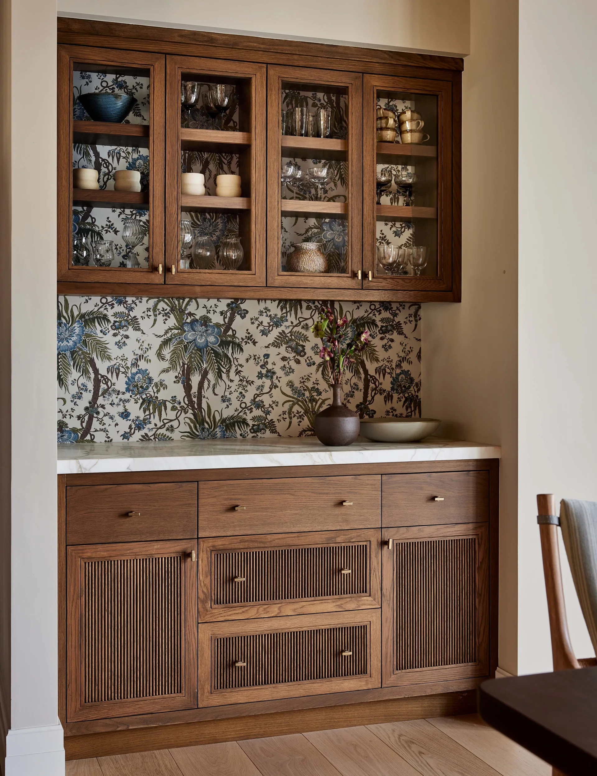 A built-in warm wood cabinet features glass-front upper doors, a white marble countertop, and blue botanical wallpaper on the backsplash.