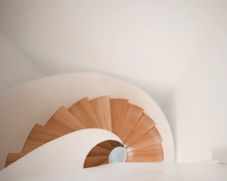 Looking down a spiral staircase featuring light wood treads and a smooth curved white railing against white walls.
