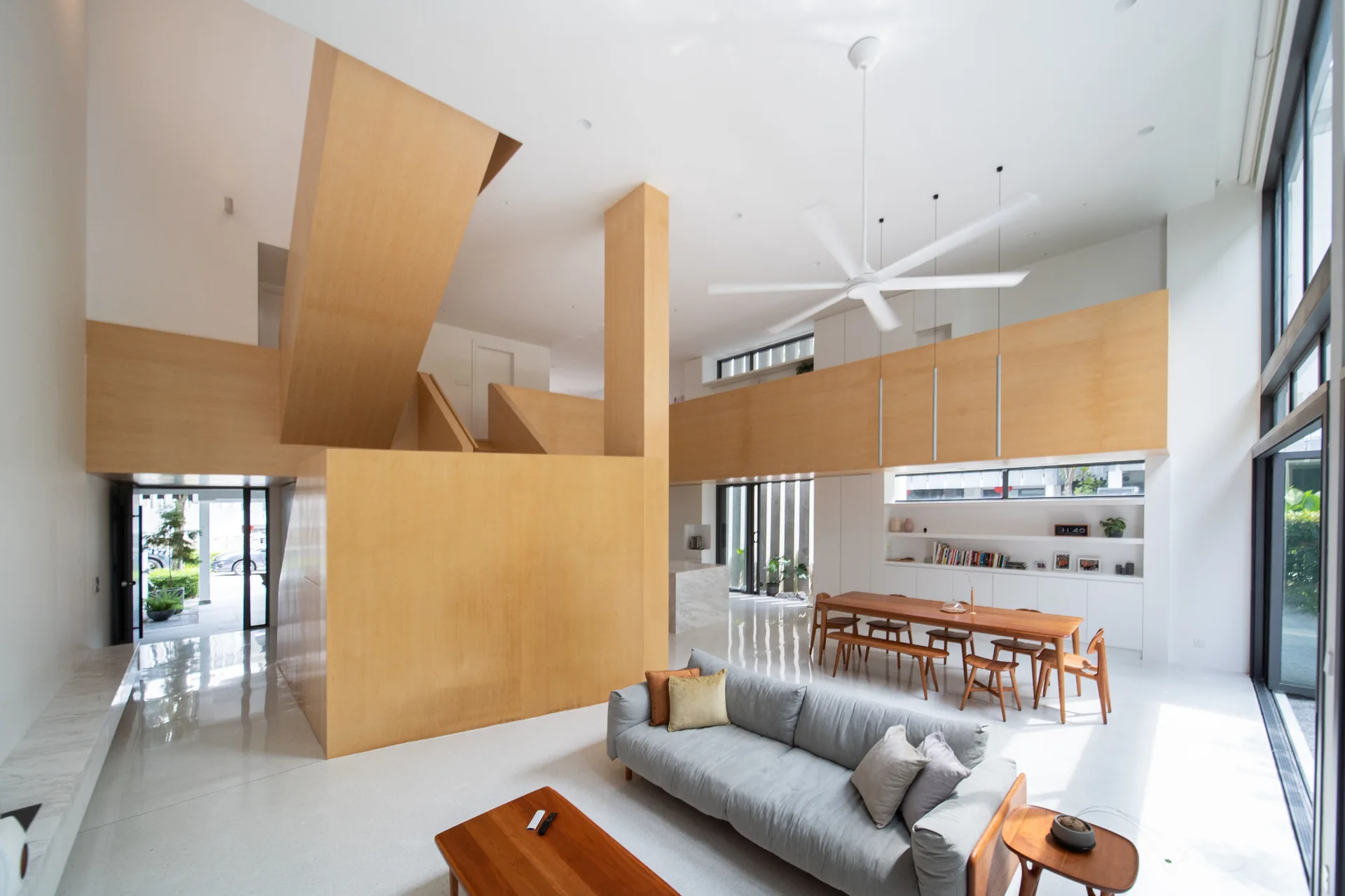 A modern open-plan living and dining area featuring a massive light wood staircase structure, a grey sofa, white floors, and a wood dining table.