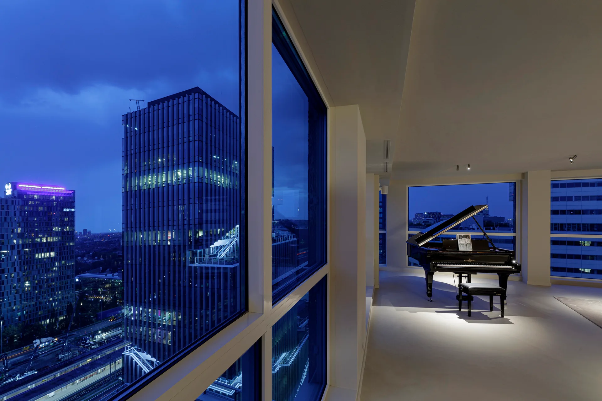 A black grand piano stands in a minimalist room with a white floor and expansive windows overlooking high-rise city buildings at dusk.