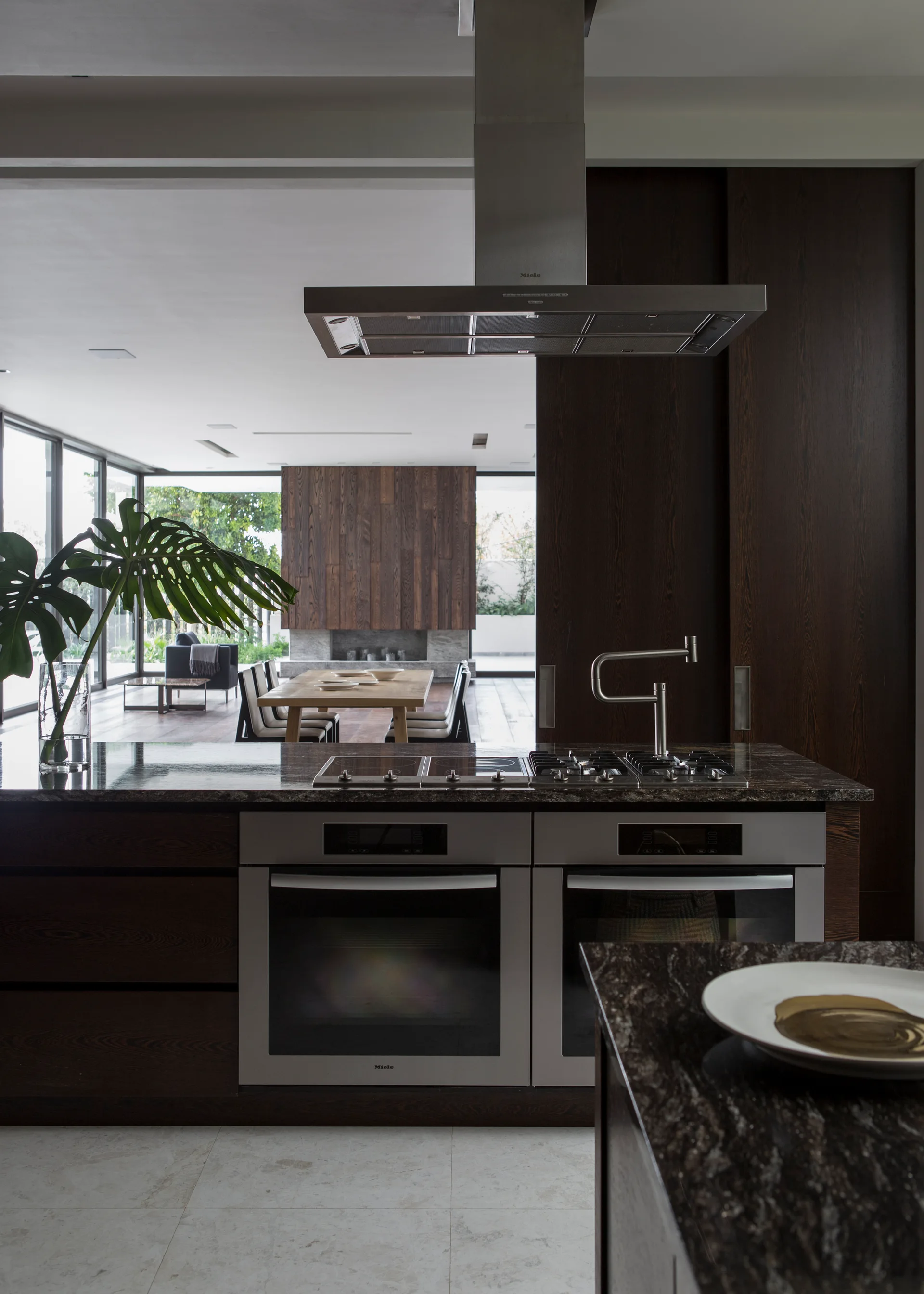 A modern kitchen island features a stone countertop, dual ovens, a cooktop, and a stainless steel range hood with a dining area in the background.