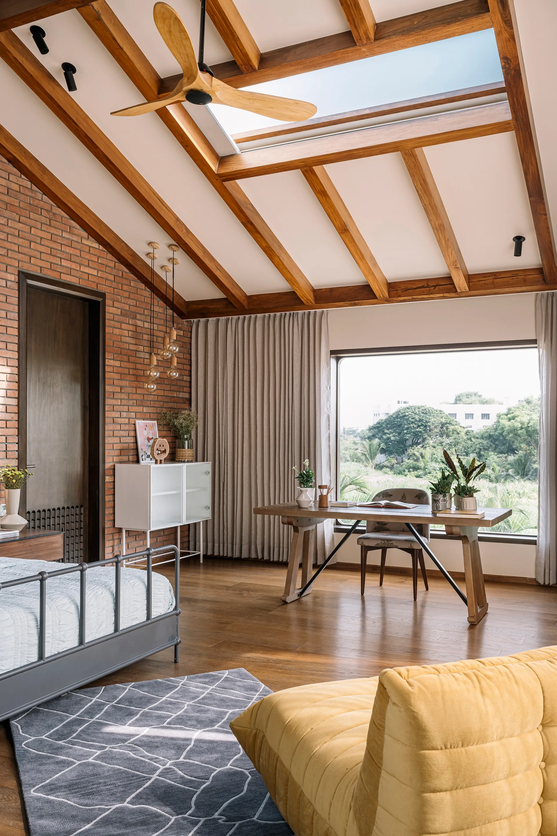 A bedroom featuring an exposed brick wall, a vaulted ceiling with wood beams and a skylight, a wooden desk near a window, and a metal bed.