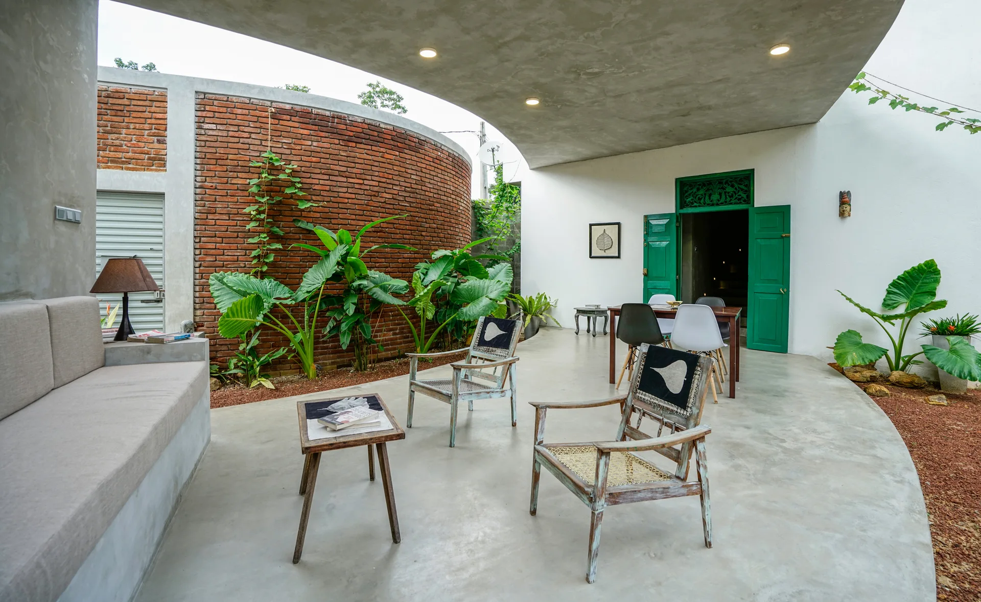 A covered concrete patio features a curved red brick wall, two distressed wooden armchairs, and lush green plants under a concrete roof overhang.