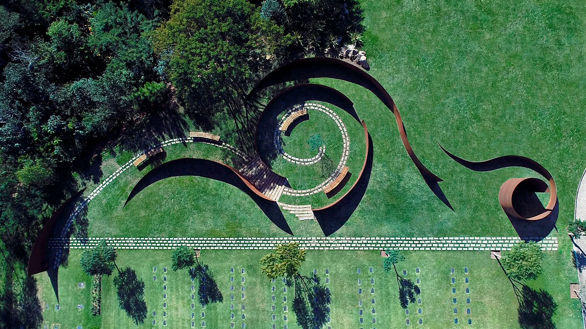 An aerial view of a manicured green lawn with a spiraling weathered steel sculpture, curved stone paths, and wooden benches.