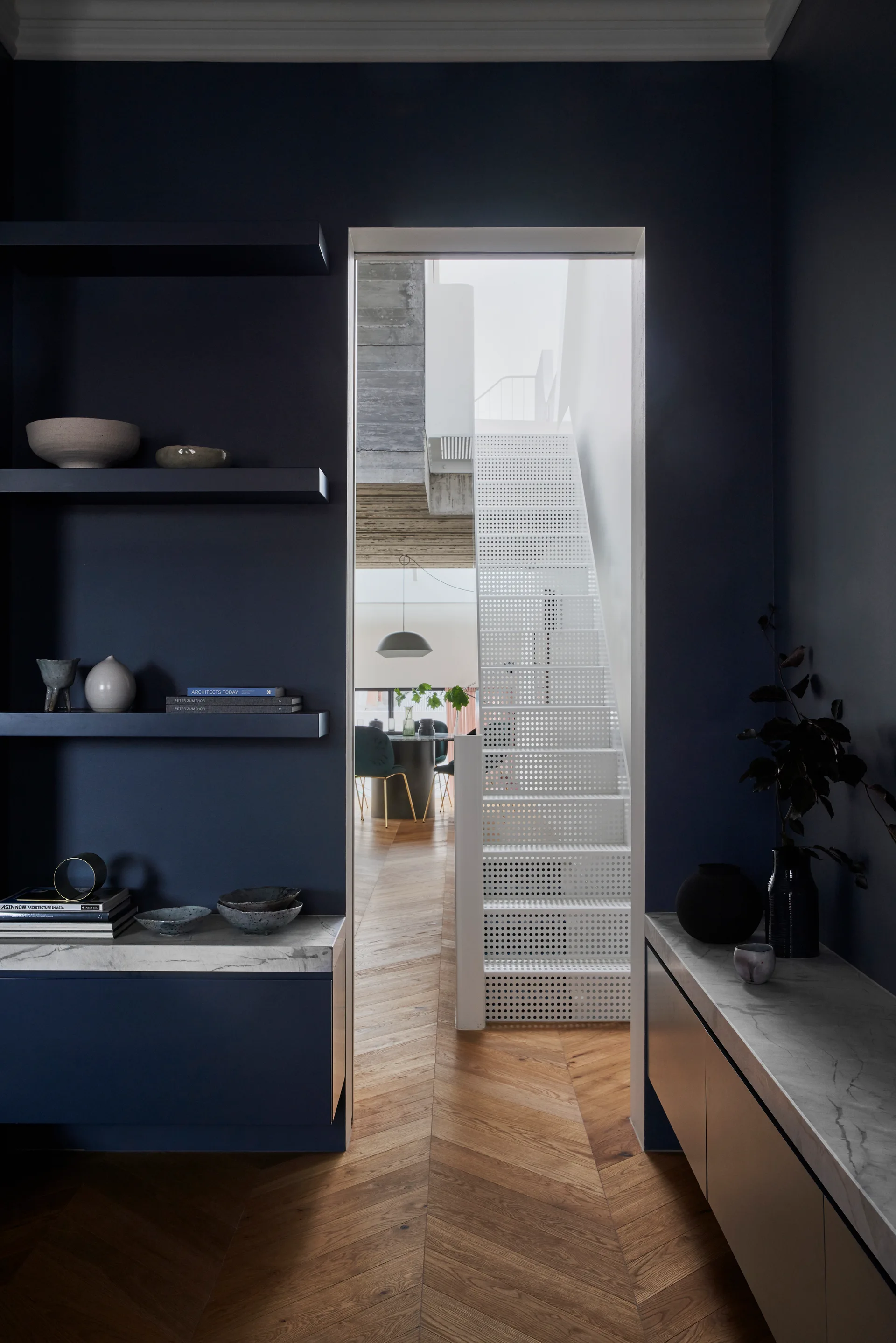 A dark navy hallway with chevron wood floors and open shelving looks through a doorway toward a white perforated metal staircase in a bright room.