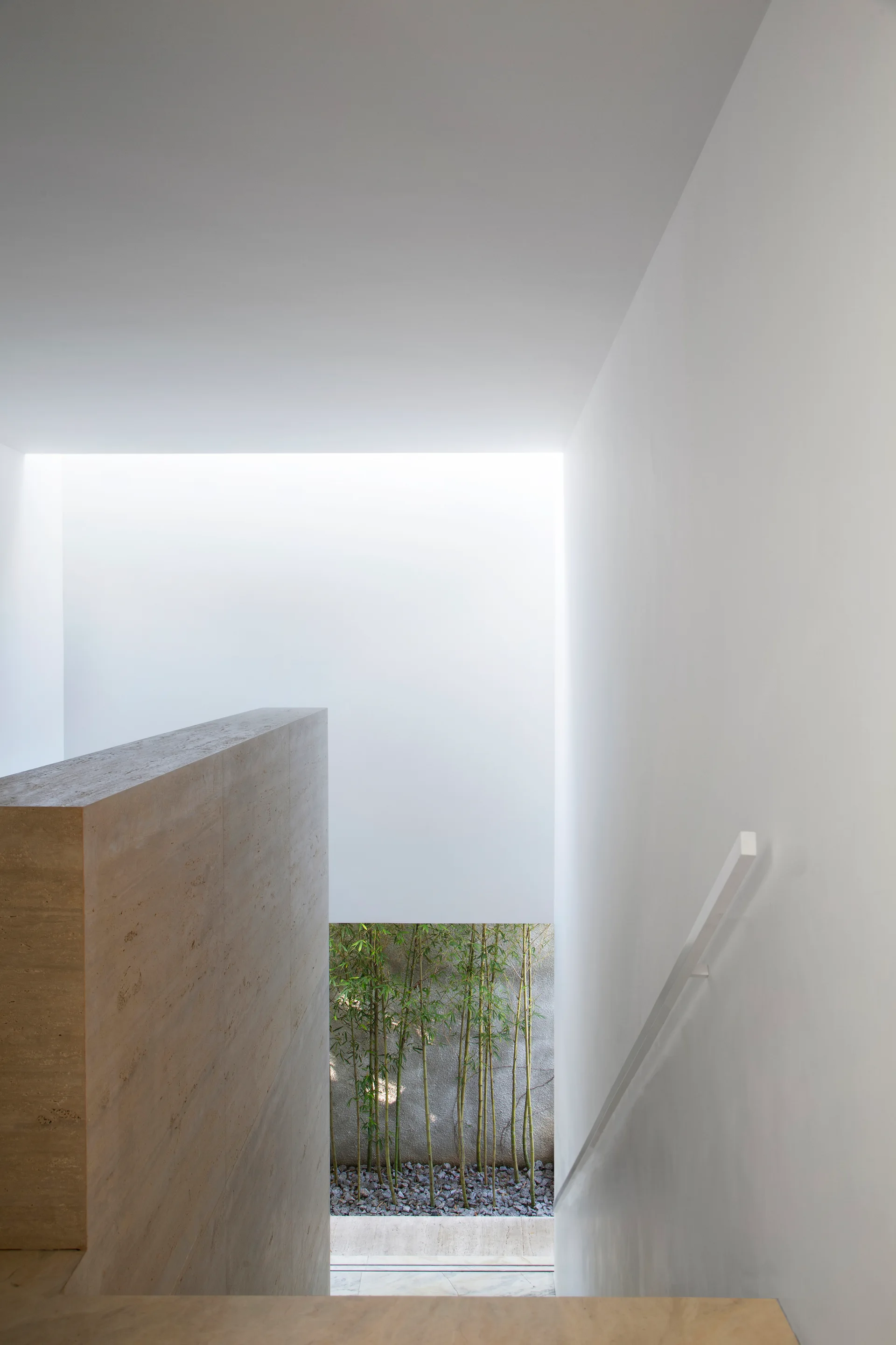 A minimalist stairwell with white walls, a textured travertine stone partition, and a low window revealing green bamboo outside.
