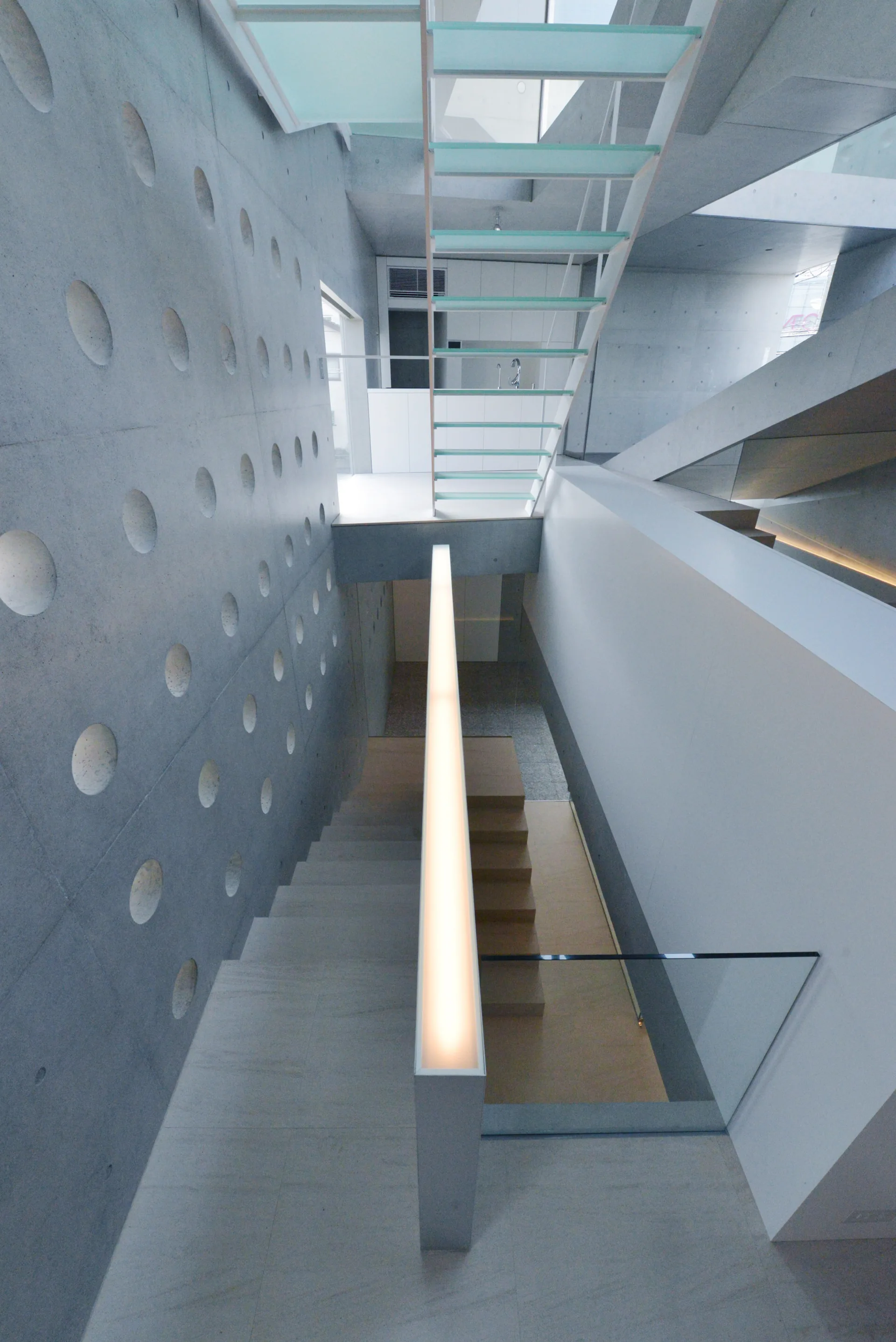 A modern multi-level staircase with translucent glass treads, an illuminated metal handrail, and a concrete wall with circular cutouts.