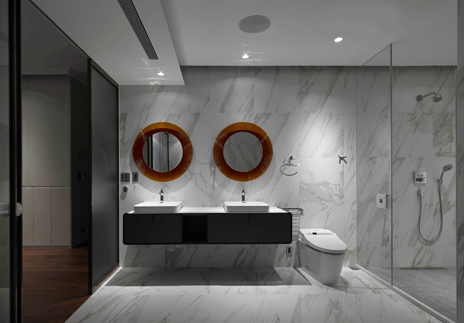 A modern bathroom features a black floating vanity, white vessel sinks, round orange mirrors, and marble tile walls.