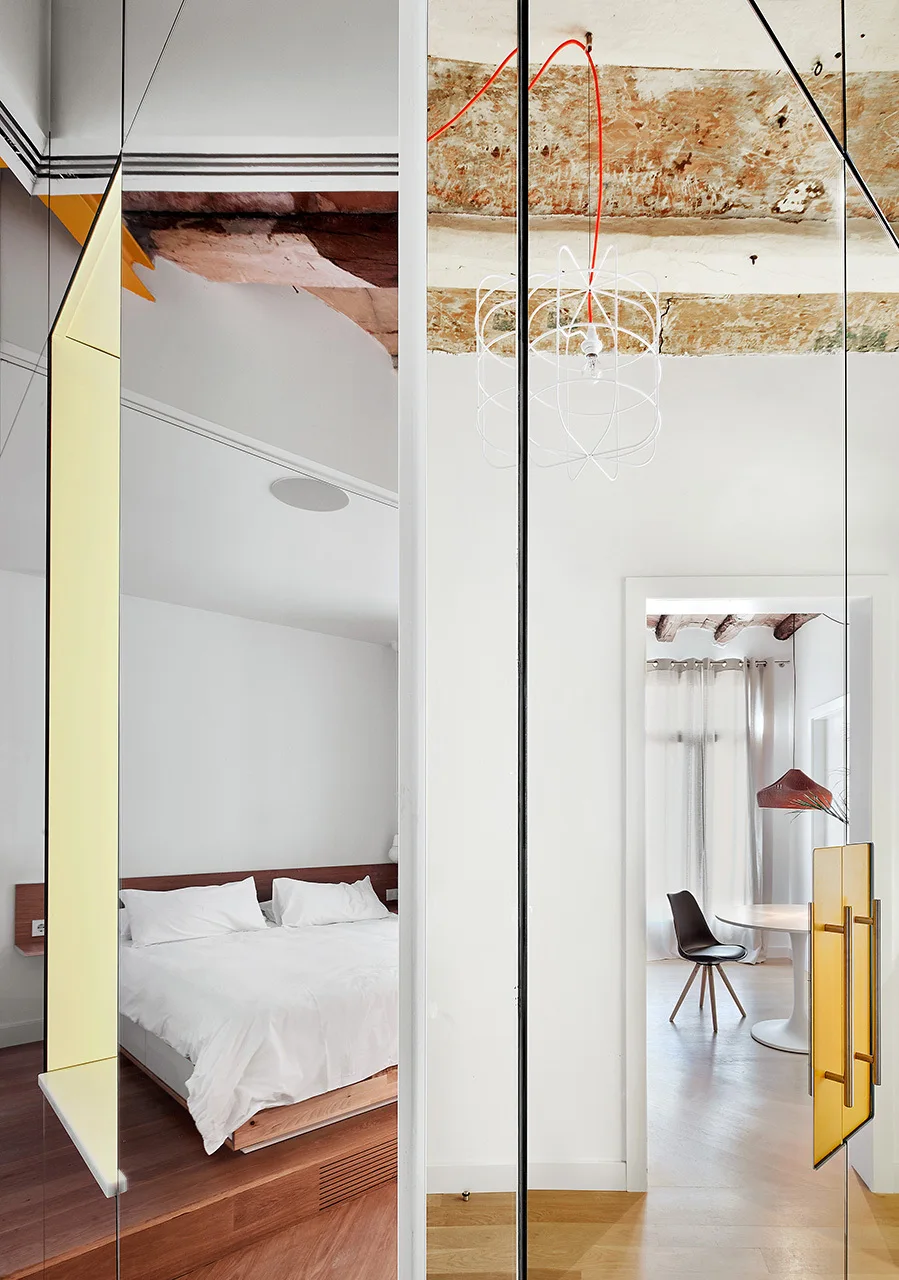 A modern bedroom featuring a platform bed is separated from a dining area by a glass partition with yellow accents under exposed wooden ceiling beams.