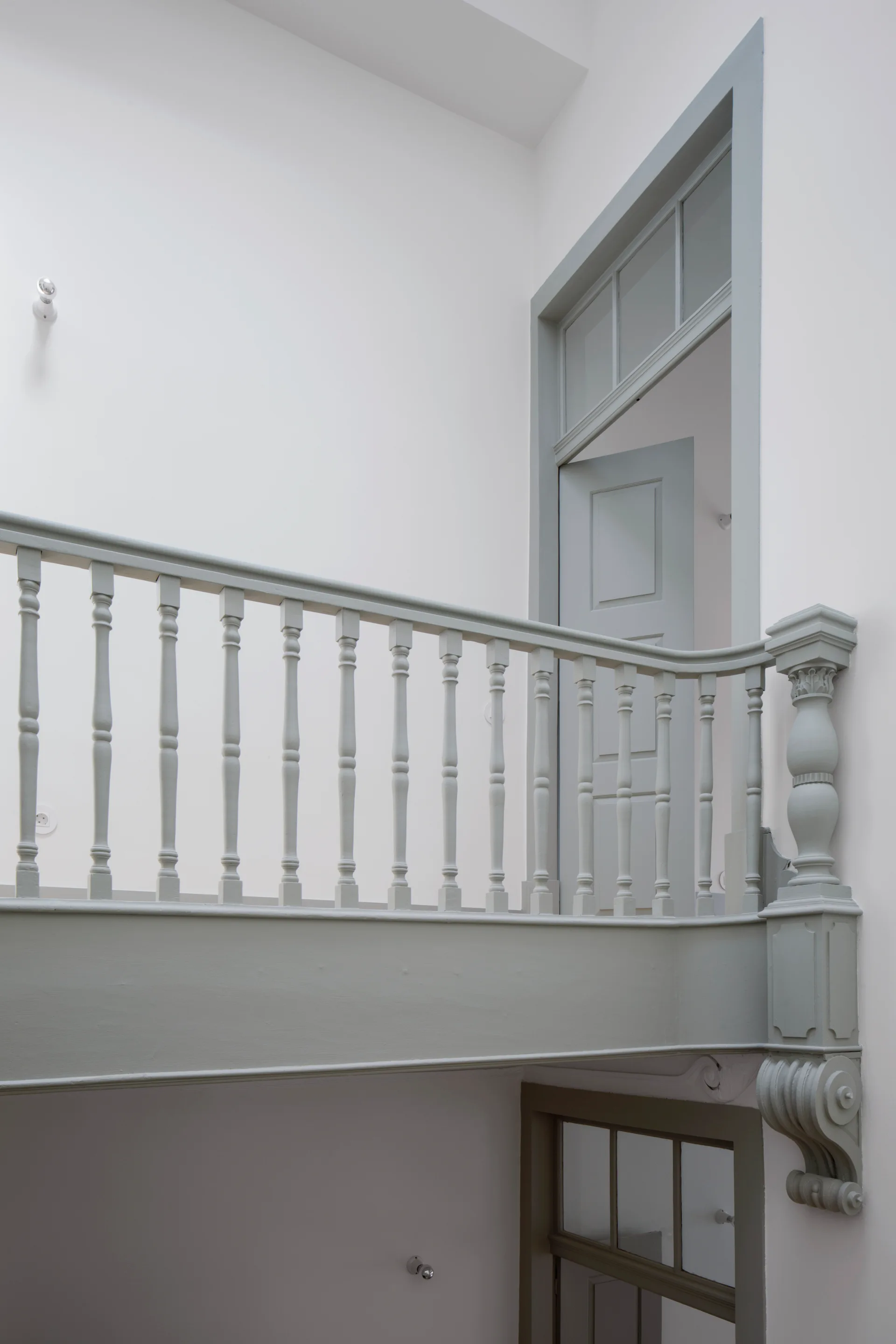 A sage grey painted wooden balustrade and matching paneled door with a transom window stand out against a minimalist white staircase landing wall.
