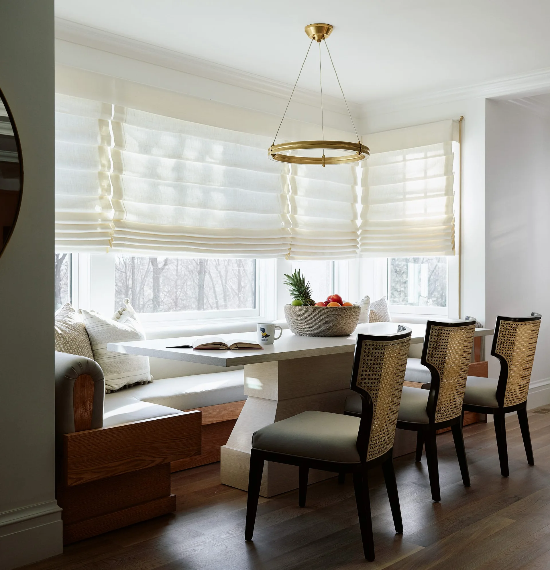 A breakfast nook in a bay window features a wooden banquette with cream cushions, a white pedestal table, three cane-back chairs, and a brass pendant light.