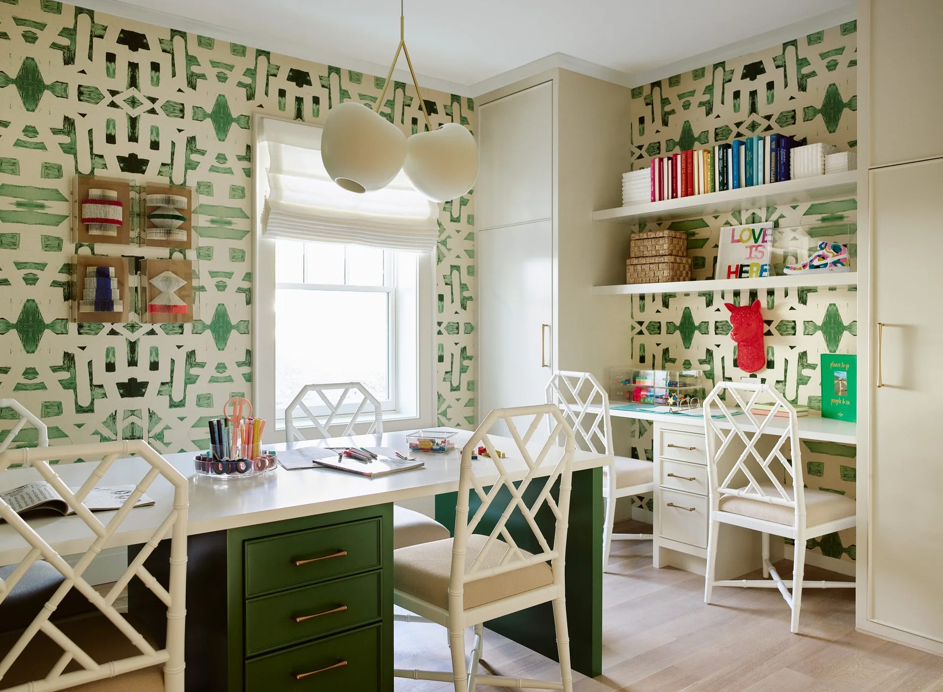A bright homework room featuring green geometric wallpaper, white lattice-back chairs, a study table with green drawers, and wall-mounted shelves.