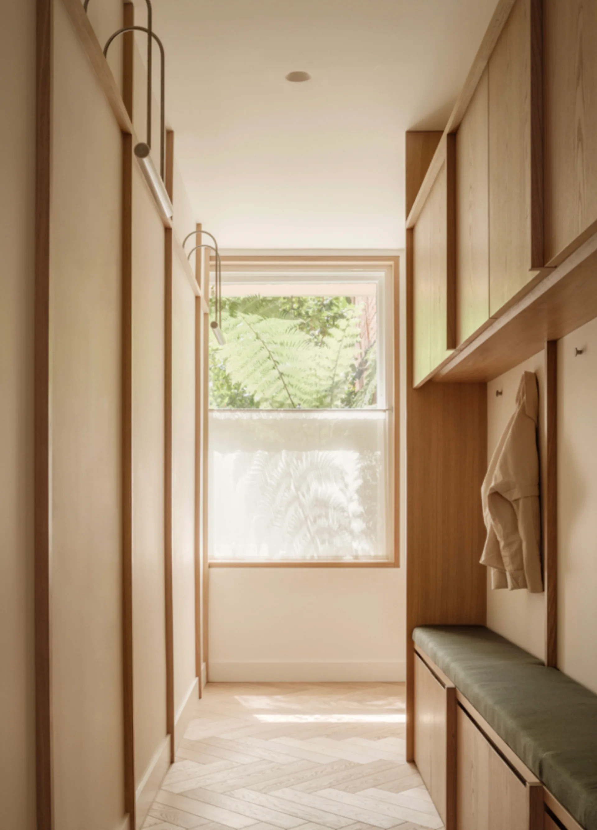 A narrow mudroom features light wood built-in cabinetry, a sage green cushioned bench, and herringbone flooring leading to a window.
