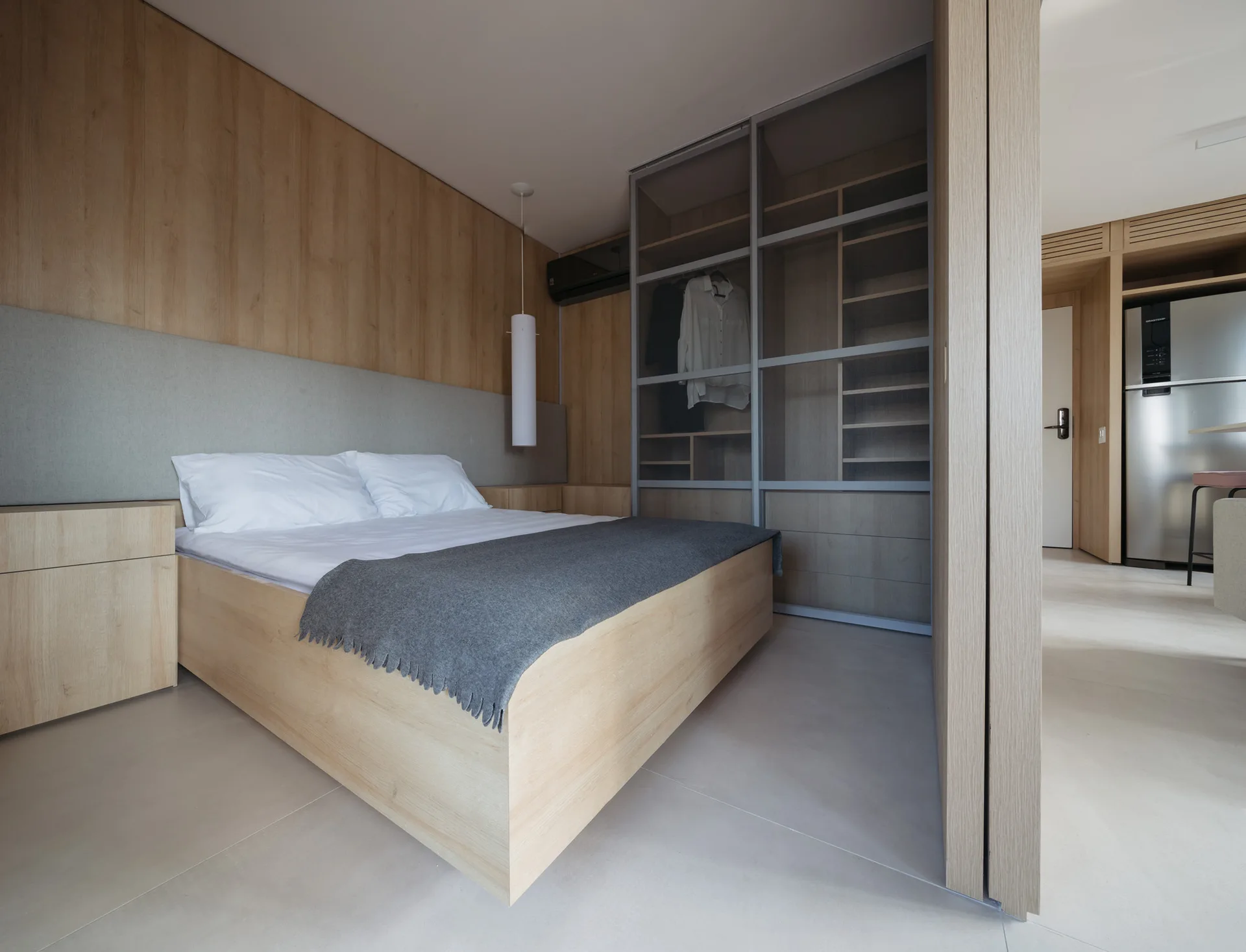 A minimalist bedroom features a light wood platform bed, matching wall paneling, an upholstered grey headboard, and a built-in wardrobe with glass doors.