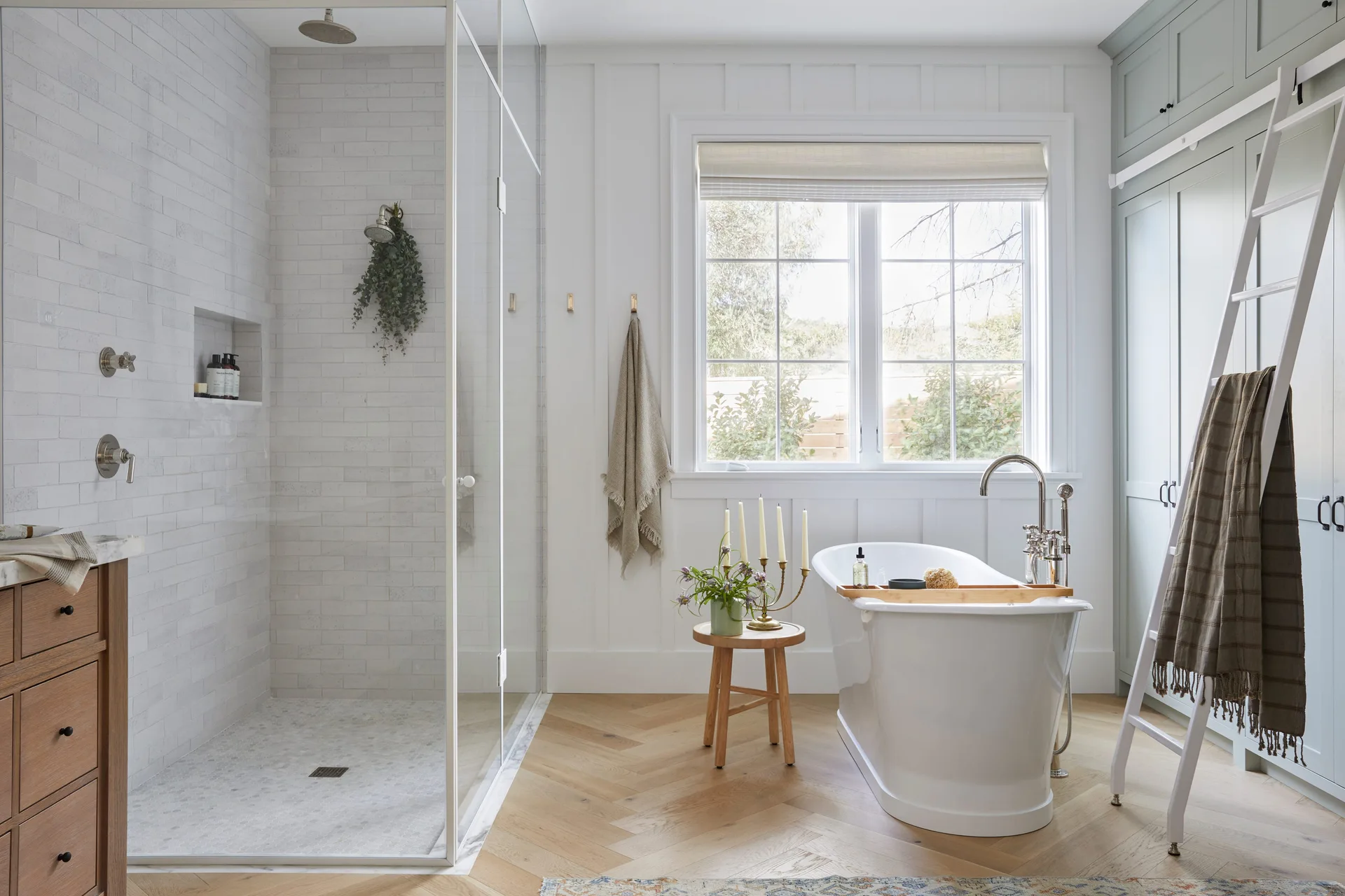 A bright bathroom featuring a white freestanding tub, a glass walk-in shower, vertical wall paneling, and light wood herringbone flooring.