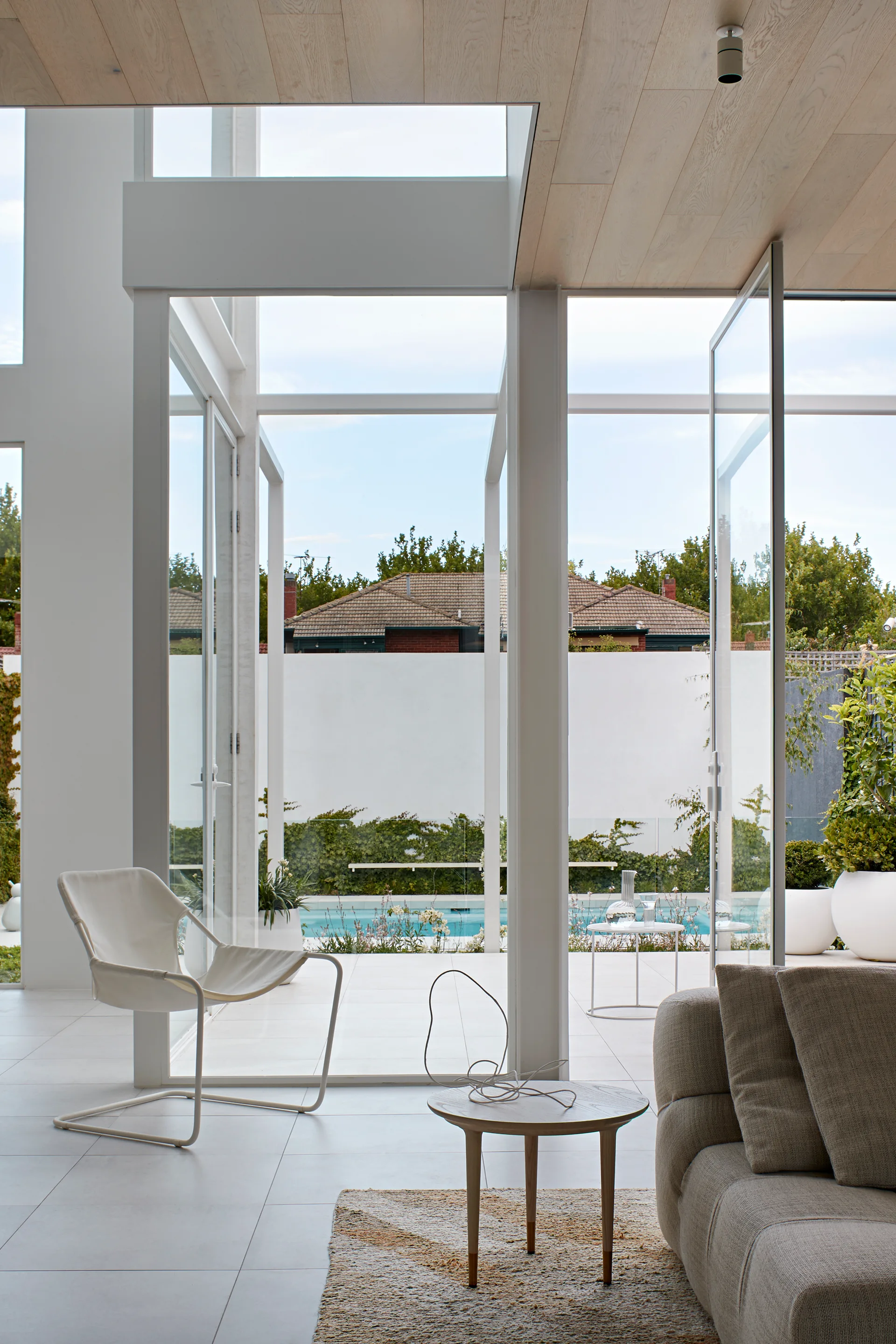 A bright living room with large glass doors opening to a swimming pool, featuring a beige sofa, a white lounge chair, and a light wood ceiling.