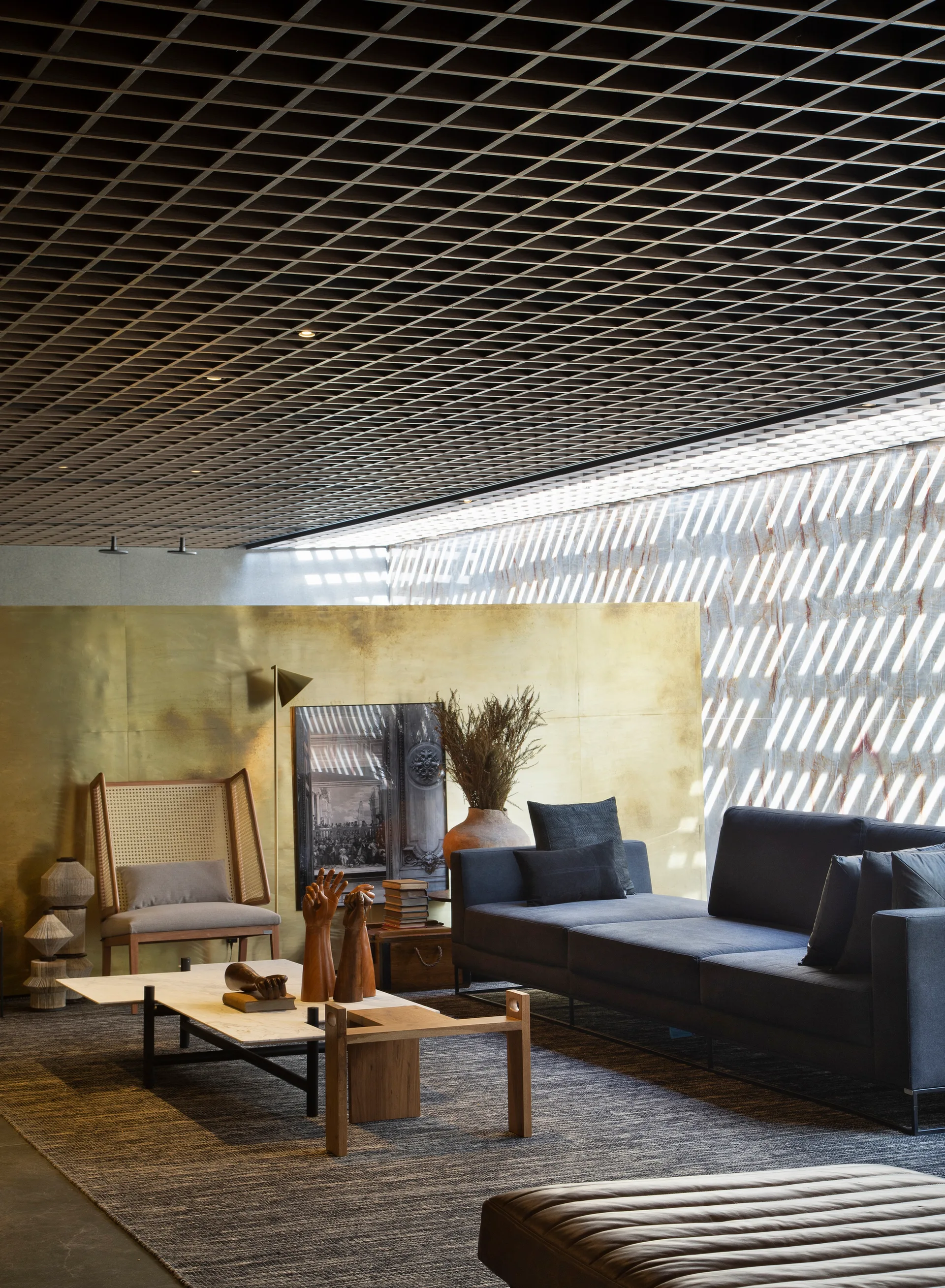 A contemporary living room features a dark grey sofa, marble-topped coffee table, high-back wicker chair, and a dark geometric grid ceiling.