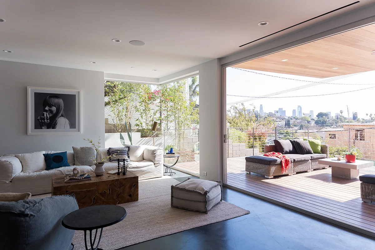 A contemporary living room with a cream sofa and block wood coffee table opens to a spacious wooden deck overlooking a city skyline.