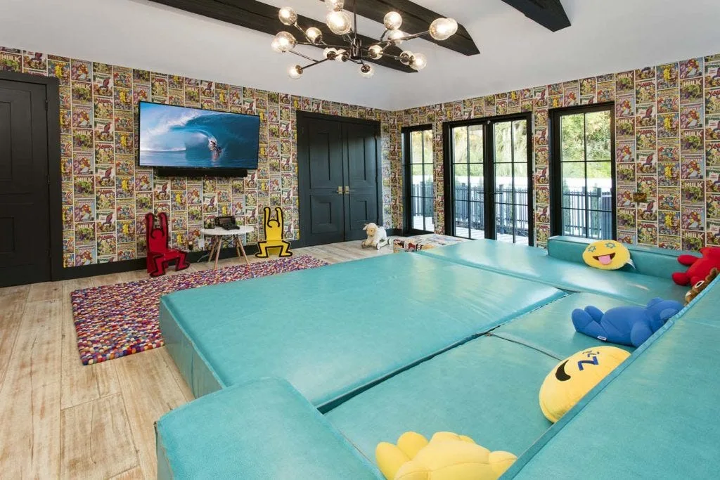 A playroom features comic book print wallpaper, a large teal modular floor sofa, dark ceiling beams, and black French doors.