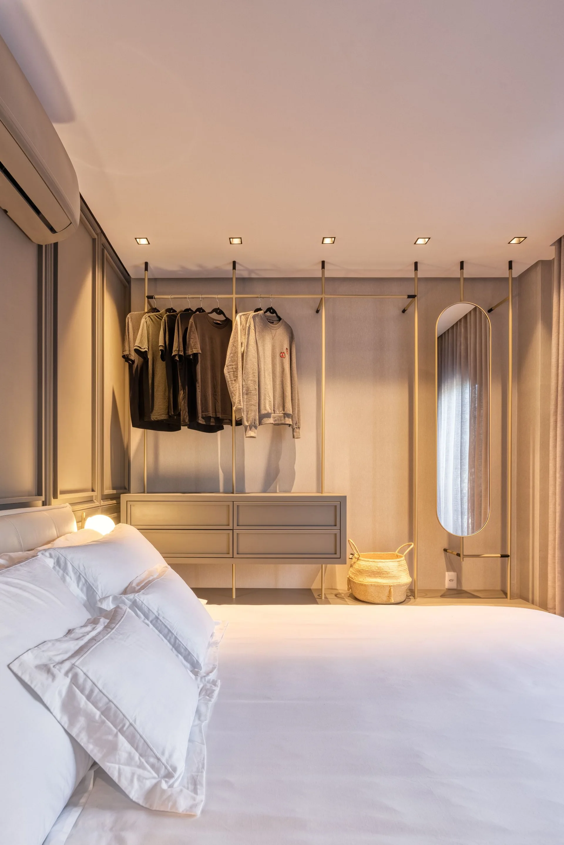 A modern bedroom featuring a bed with white pillows and an open metal closet system with drawers, hanging clothes, and an oval mirror.