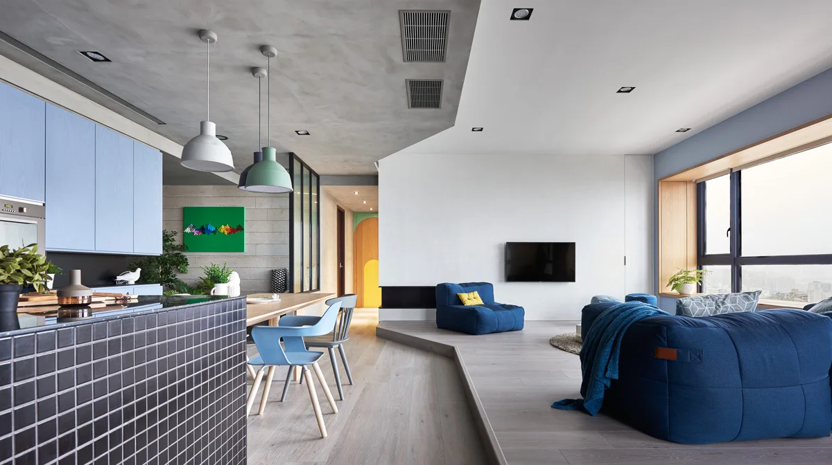 An open-plan apartment features a raised platform living room with blue modular sofas adjacent to a dining area and light blue kitchen cabinetry.