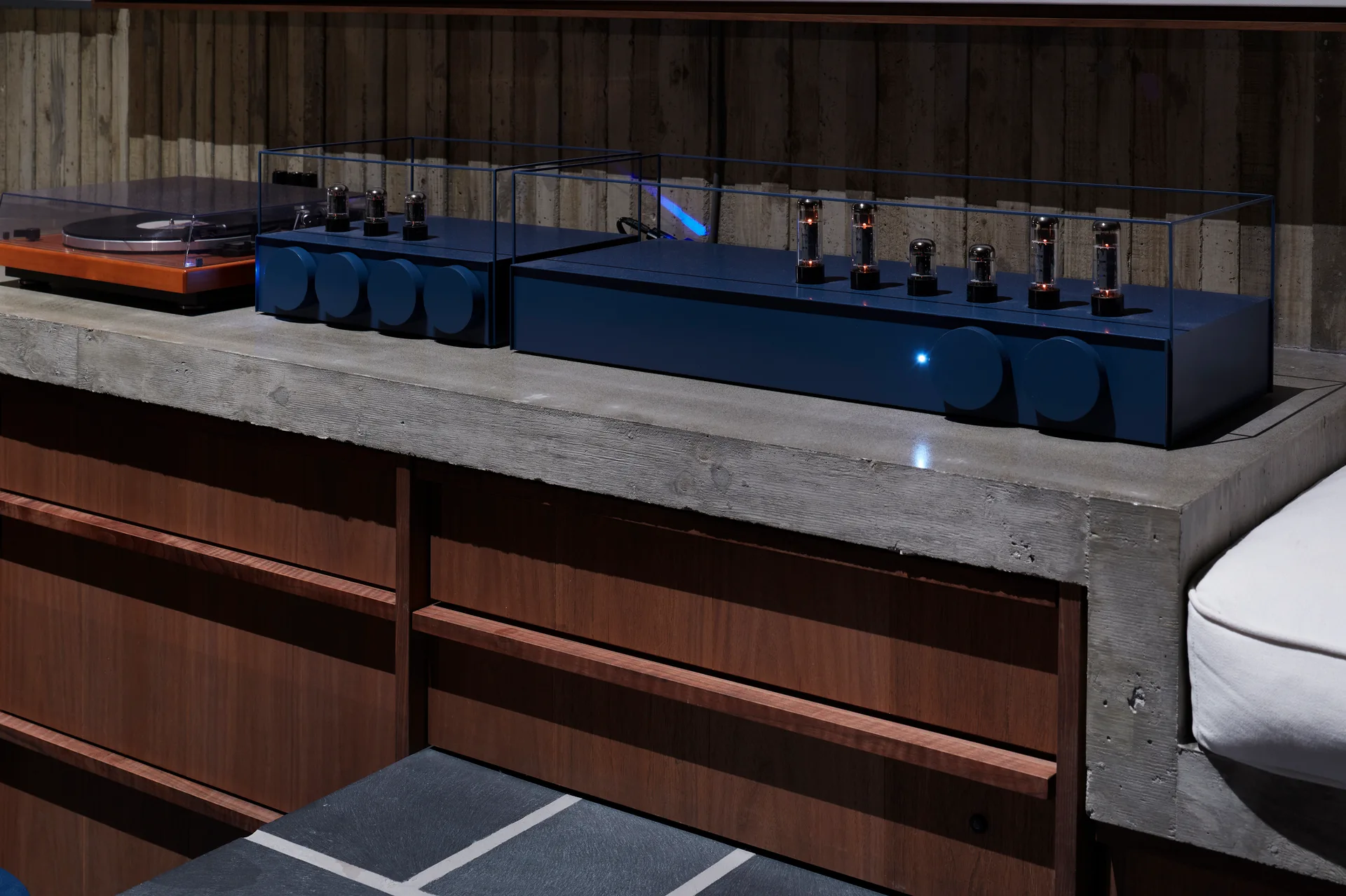 A deep blue tube amplifier and a turntable sit on a raw concrete countertop over warm wood cabinets against a vertical wood slat wall.