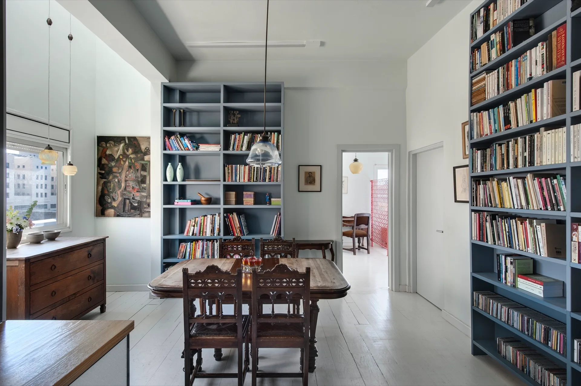 A dining area featuring an ornate wood table and chairs surrounded by large grey built-in bookshelves on a white painted plank floor.