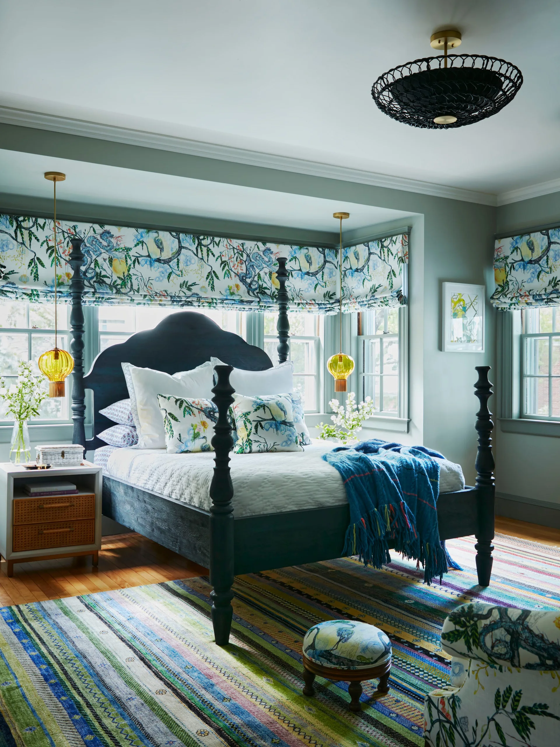 A bedroom features a black wooden four-poster bed framed by botanical-print roman shades, amber glass pendant lights, and a colorful striped area rug.