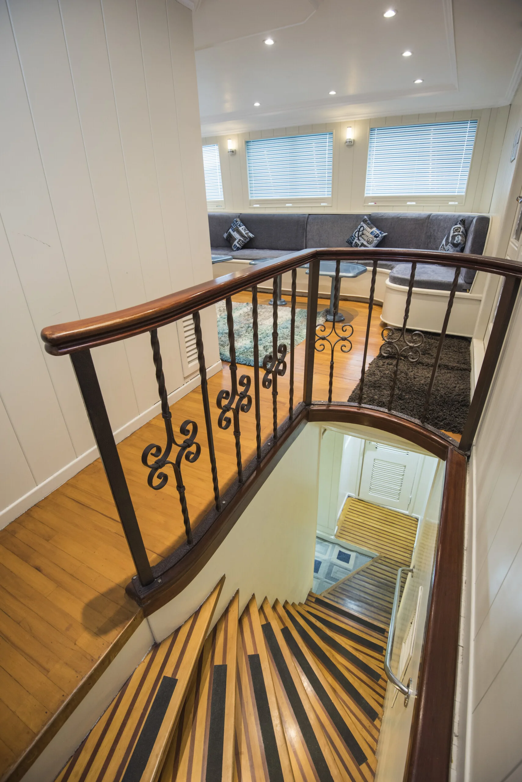 A varnished wood staircase with striped black treads leads down past a curved wrought-iron railing into a compact lounge with a gray built-in sofa.