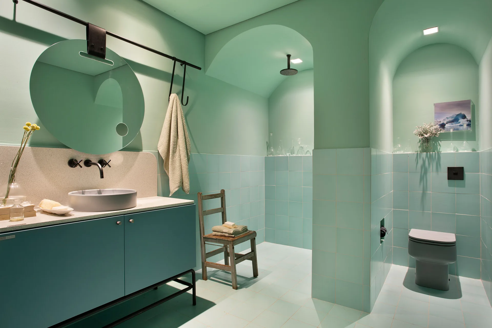 A monochromatic mint green bathroom features a round mirror, blue vanity, square-tiled walls, and two arched alcoves for the shower and toilet.