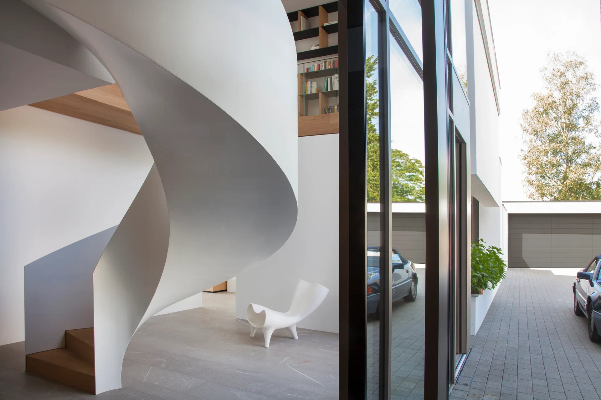 A minimalist foyer features a large white sculptural spiral staircase with wood treads next to floor-to-ceiling glass windows.