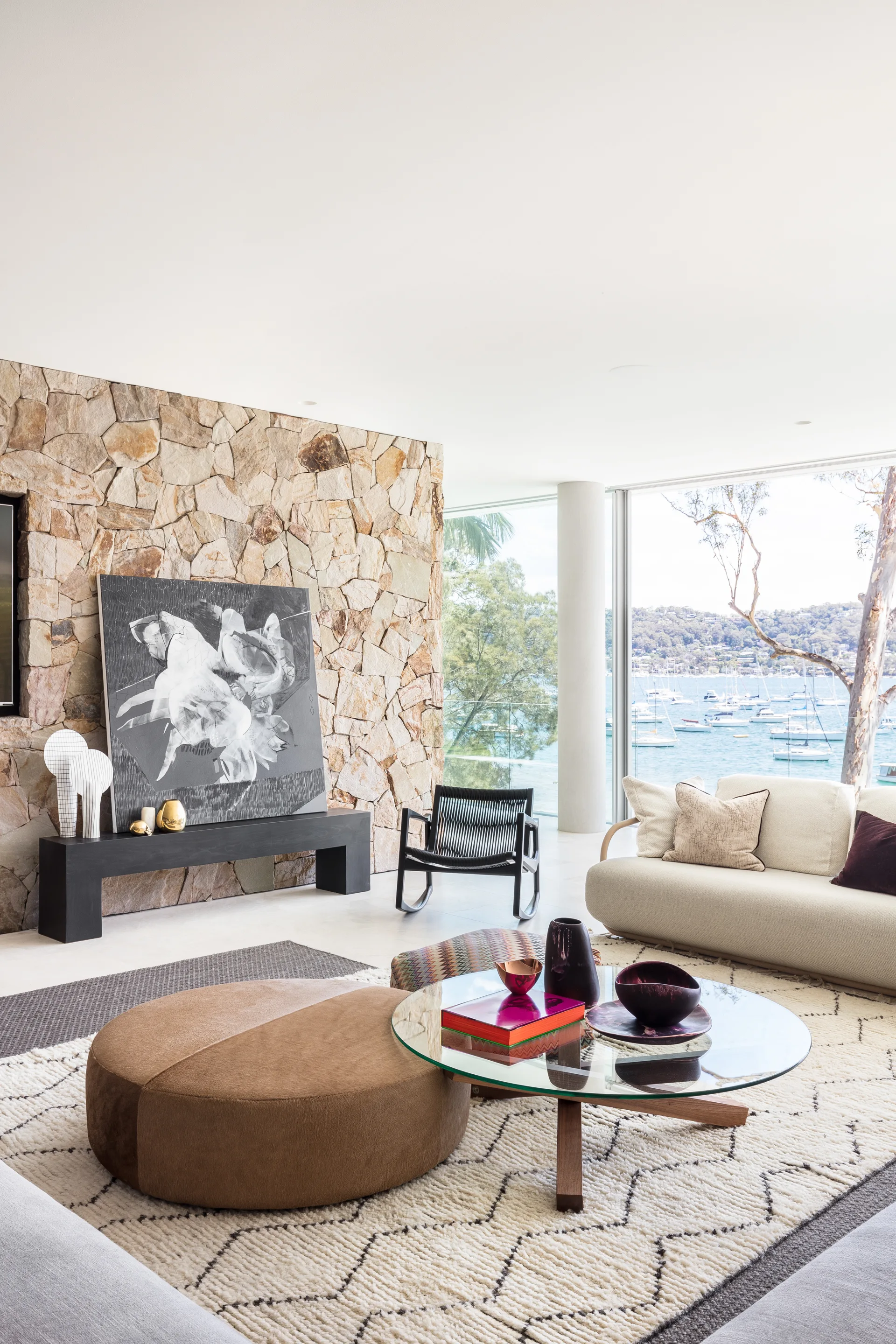 A modern living room features a textured stone accent wall, a curved cream sofa, a glass coffee table, and large windows overlooking the water.