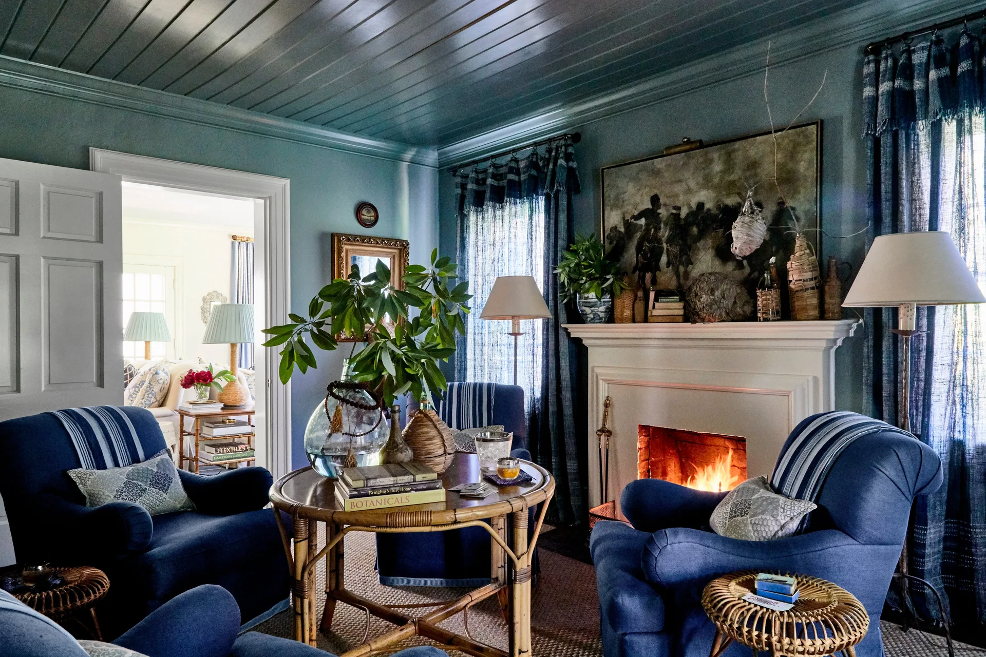 A cozy blue living room features matching upholstered armchairs, a lit white fireplace, and a round rattan coffee table beneath a painted plank ceiling.