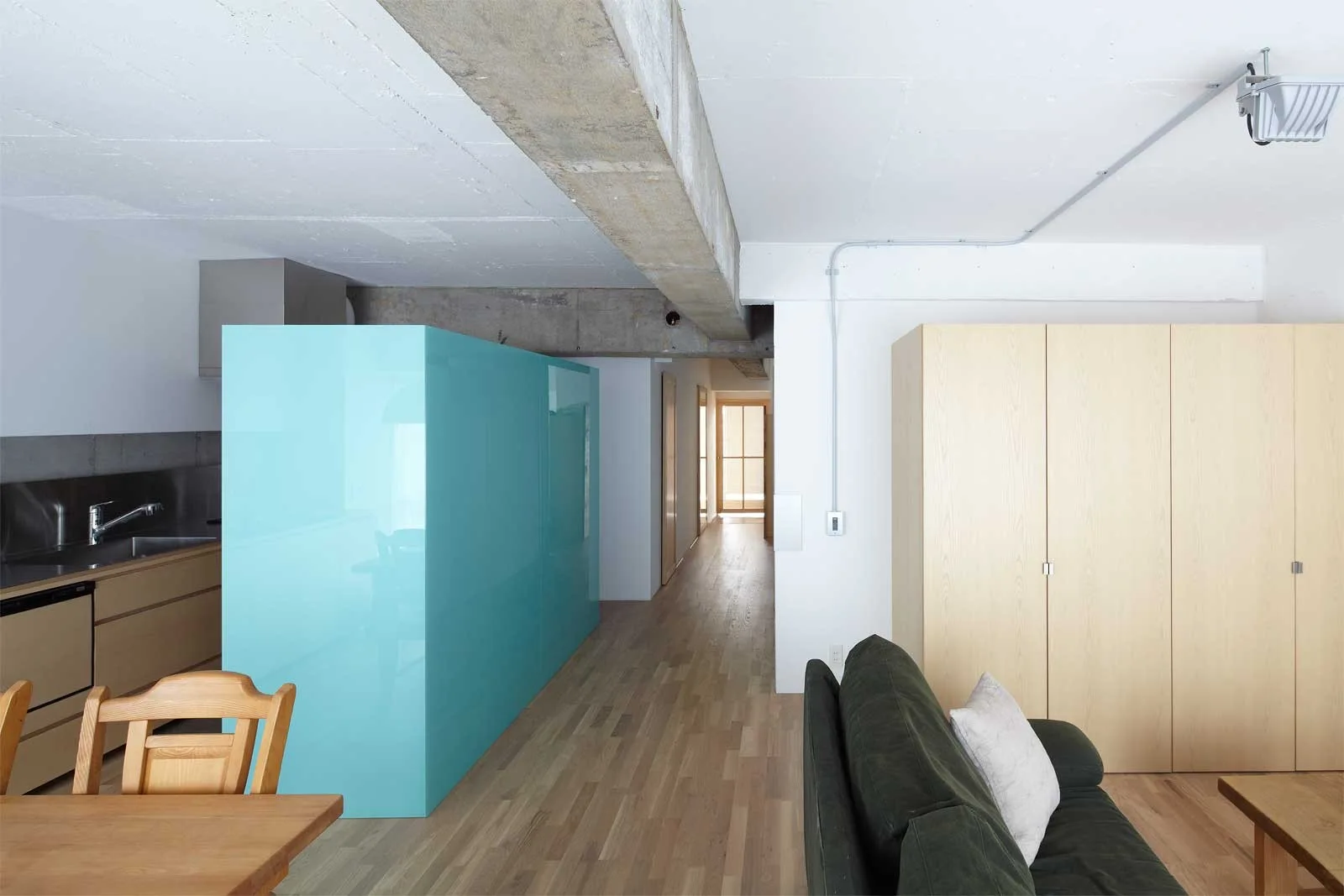 An open-plan living area and kitchen feature a glossy turquoise room divider, exposed concrete ceiling beams, wood flooring, and a dark green sofa.