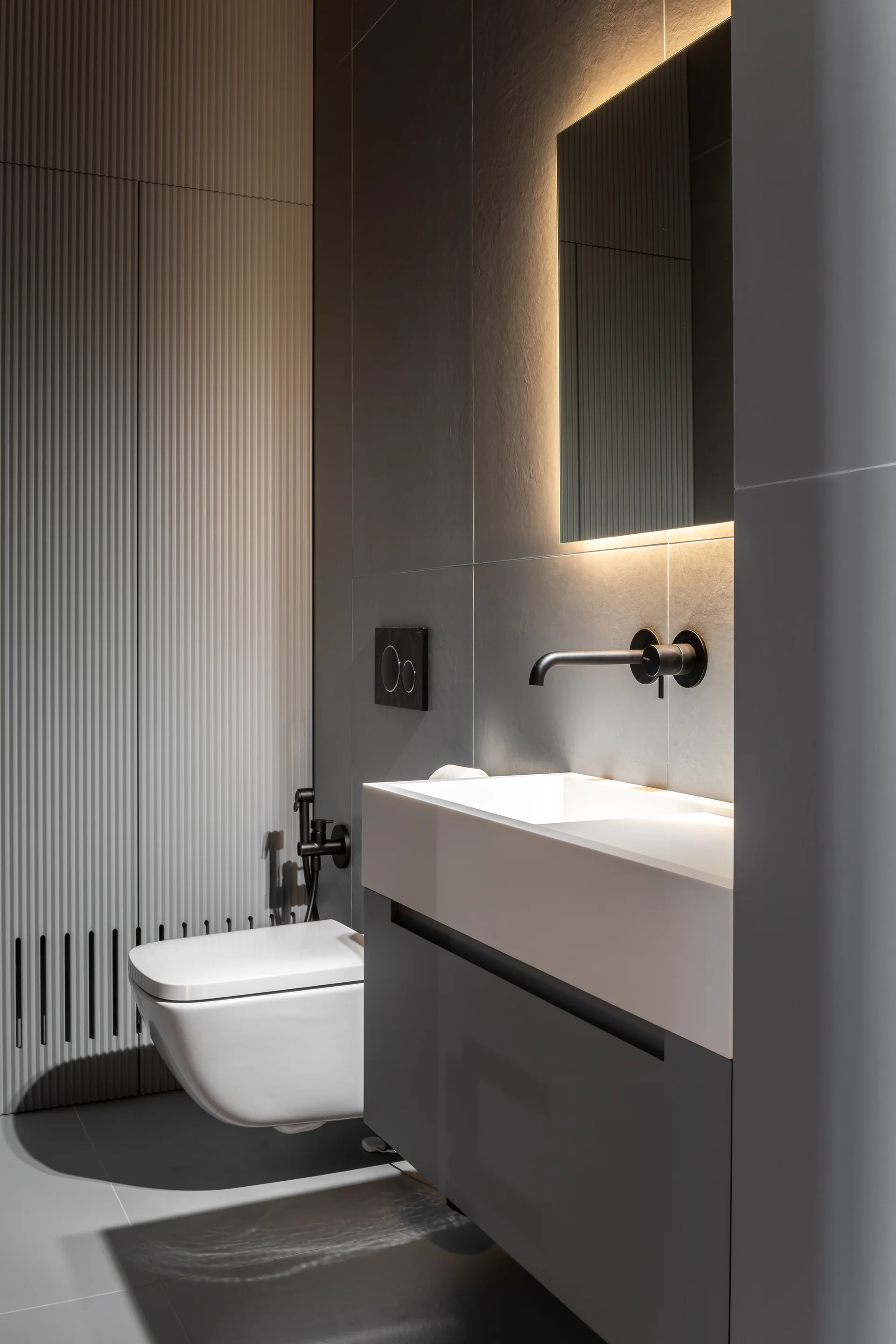 A modern grey bathroom featuring a floating white sink vanity, a wall-mounted toilet, matte black fixtures, and a rectangular backlit mirror.