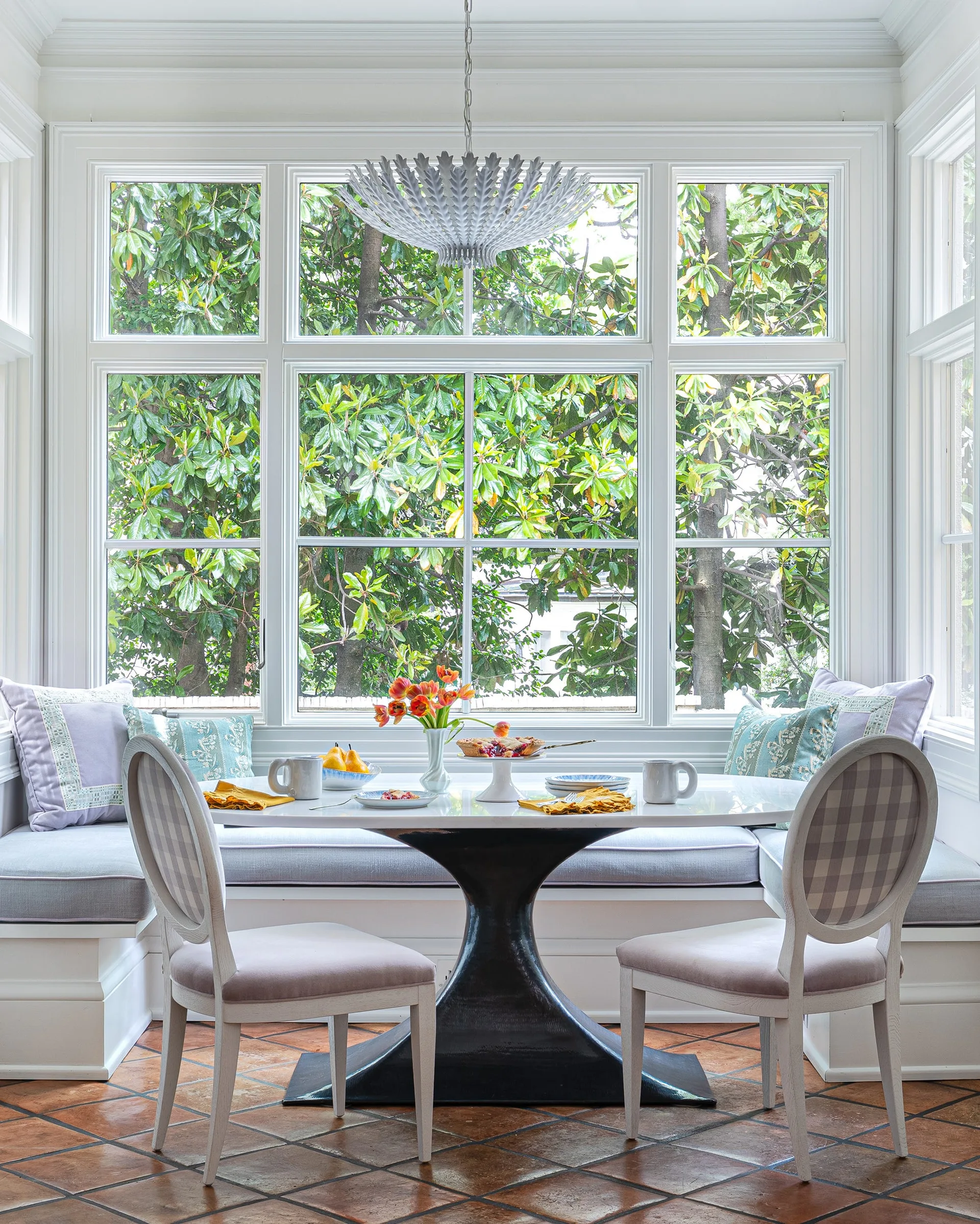 A bright breakfast nook features a white U-shaped banquette, an oval pedestal table, and checkered chairs set against large windows and a terracotta floor.