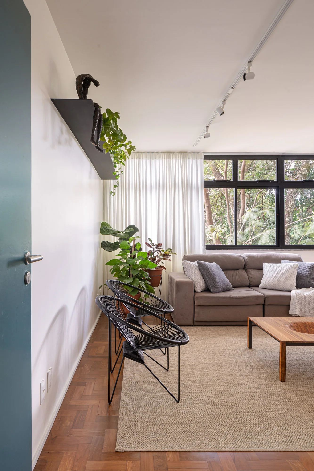 A bright living room features parquet wood floors, a grey sofa by a window with sheer curtains, and two black corded chairs.