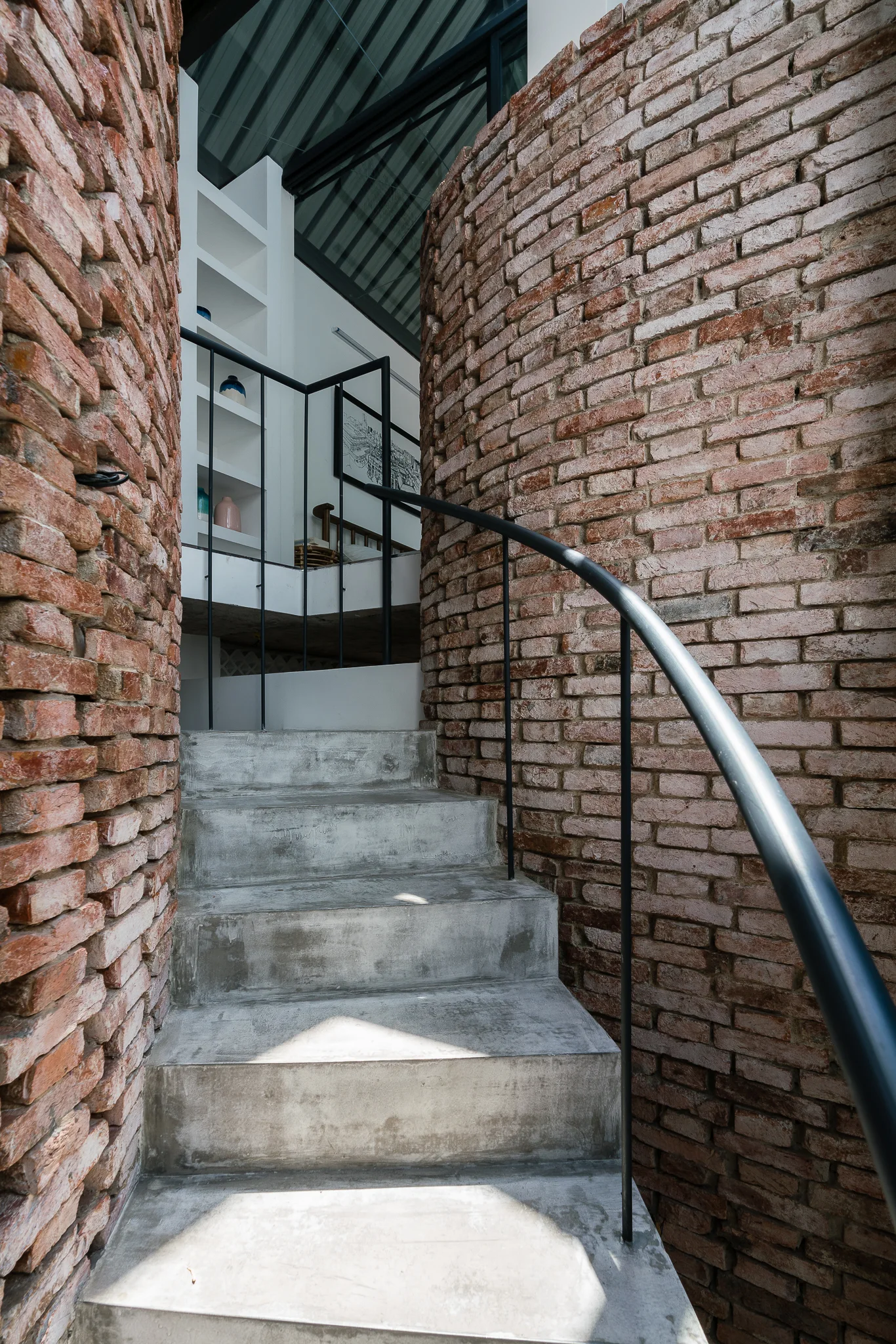 A concrete staircase curves upwards between two textured walls of exposed red brick, featuring a slim black metal handrail leading to a lit landing.