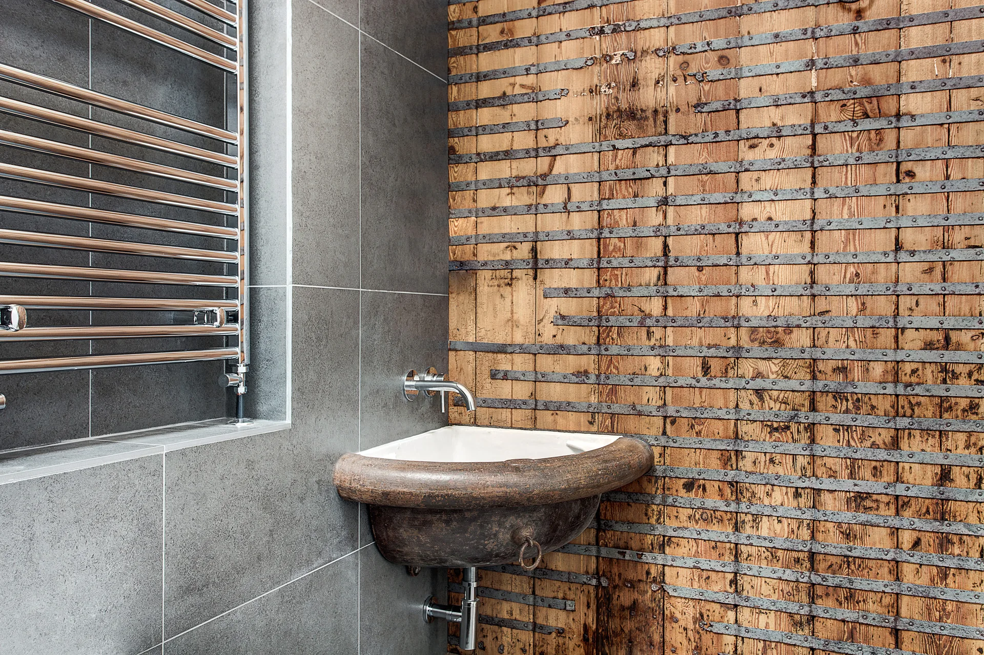 A wall-mounted sculptural sink rests against a rustic wood-planked wall with metal straps next to a gray tiled wall with a heated towel rack.