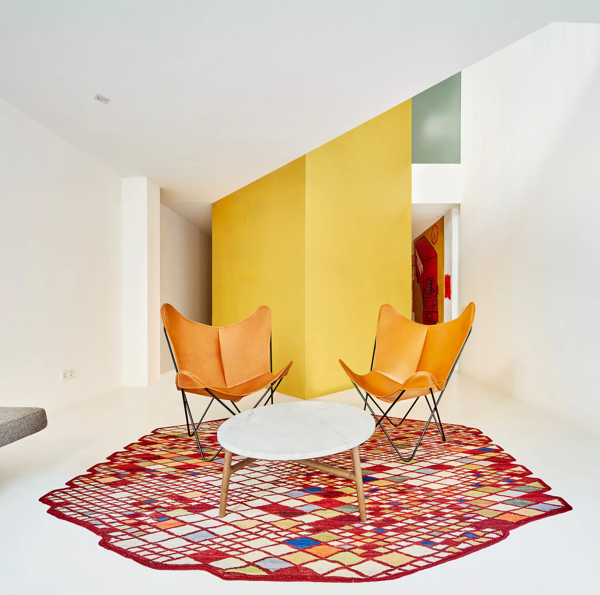 Two tan leather butterfly chairs sit around a small marble coffee table on a colorful geometric rug in front of a bright yellow wall.