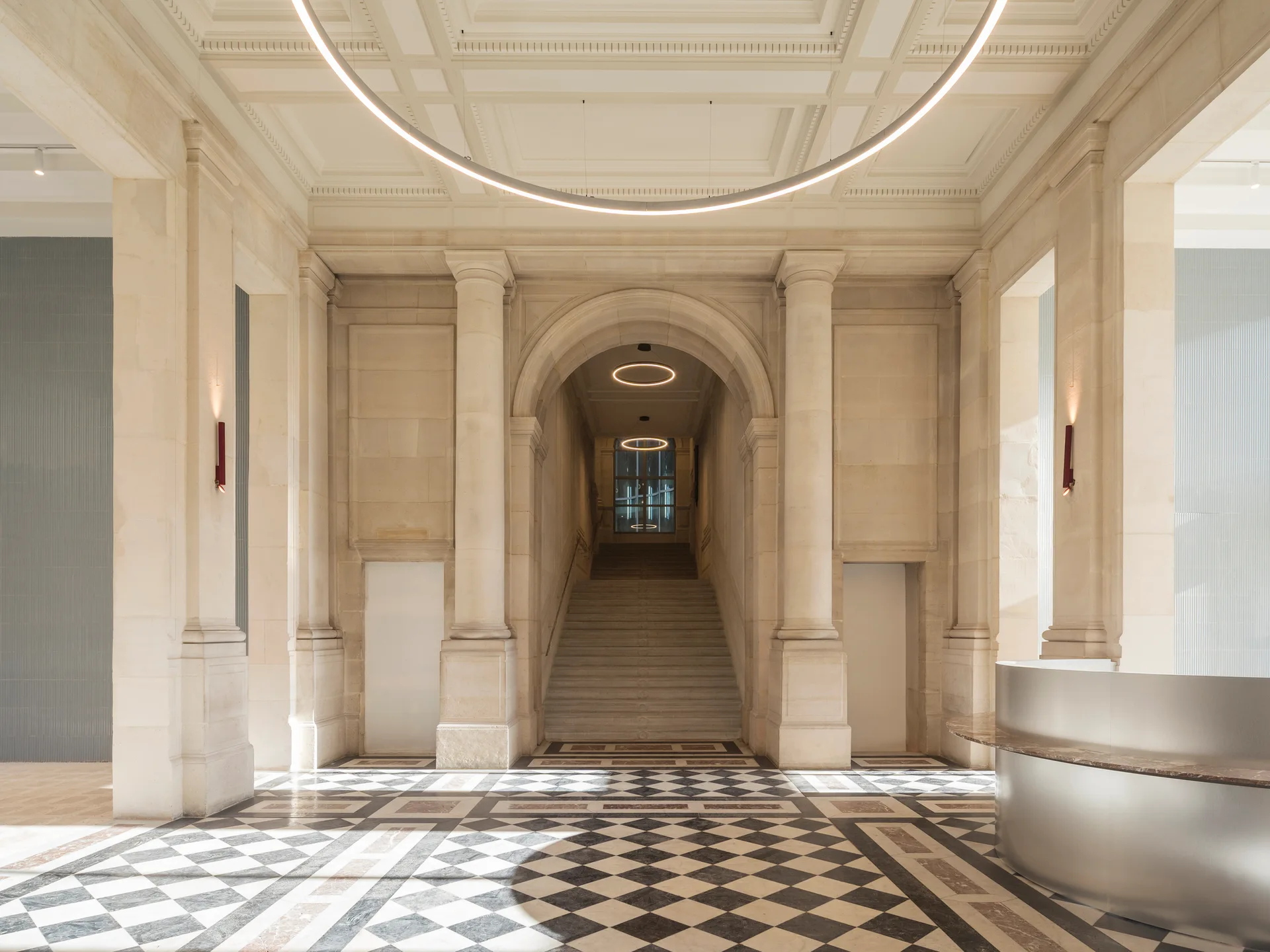A grand stone entry hall features a black and white checkerboard floor, neoclassical columns, a central staircase, and a large circular light fixture.