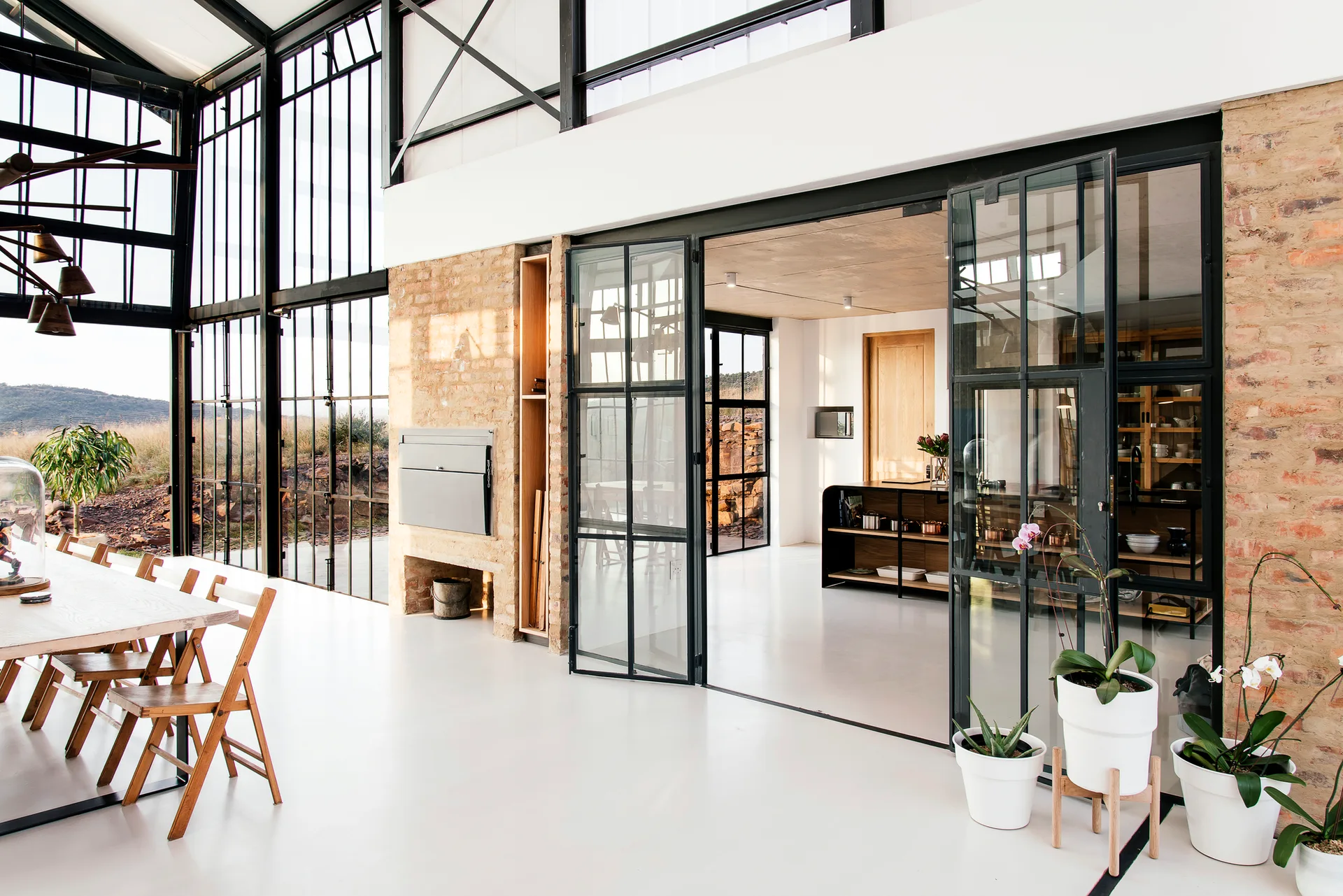 A bright industrial dining room features a wooden table with folding chairs next to an exposed brick wall and large black steel-framed folding glass doors.