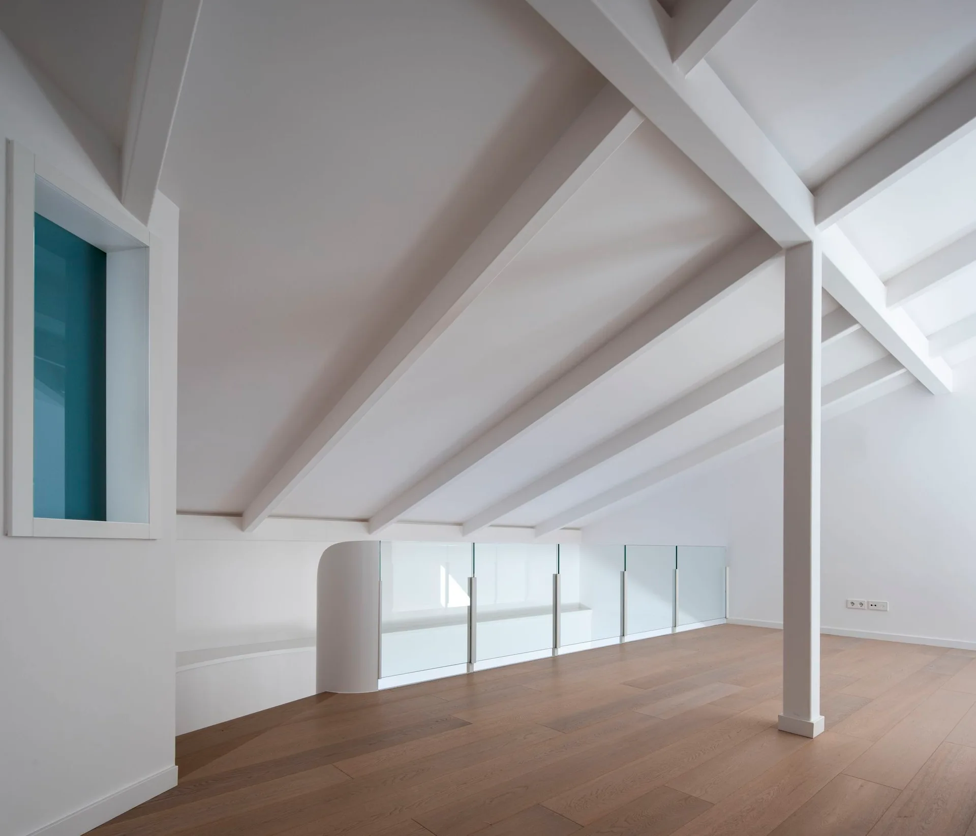 A minimalist attic loft space features a sloped ceiling with painted white structural beams, light wood plank floors, and a clear glass safety railing.