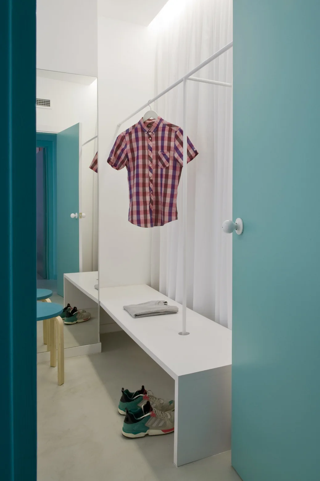 A minimalist dressing room features a white console bench under a thin hanging rod, next to bold teal doors and a full-length wall mirror.