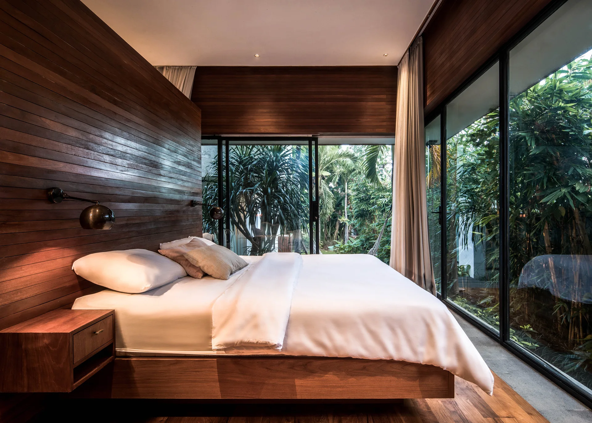A modern bedroom features a wood platform bed and matching wood-clad walls next to large glass doors opening to a lush garden.